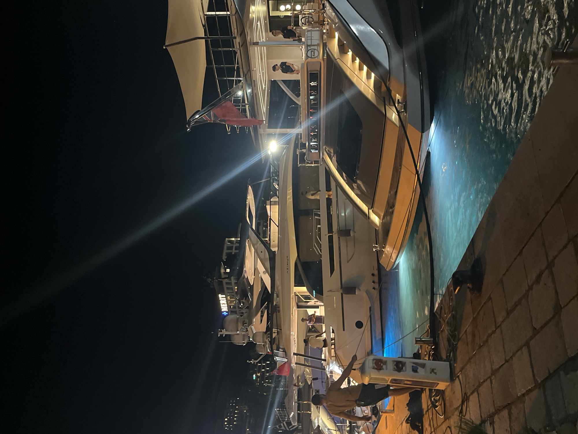 This is a night scene in a marina in Budva, Montenegro, featuring several docked luxury yachts. In the foreground, a man wearing dark shorts and no shirt stands on a paved walkway, holding a rope connected to a white power pedestal. The water in the marina glows with an artificial blue light. Several large yachts are visible, with the one closest to the viewer featuring a sleek, modern design and illuminated interior. Another yacht behind it displays the name "AZIMUT" and has a red British flag flying from its stern. Further in the background, buildings are illuminated, suggesting a city skyline. The overall atmosphere is one of a sophisticated night out in a coastal city.