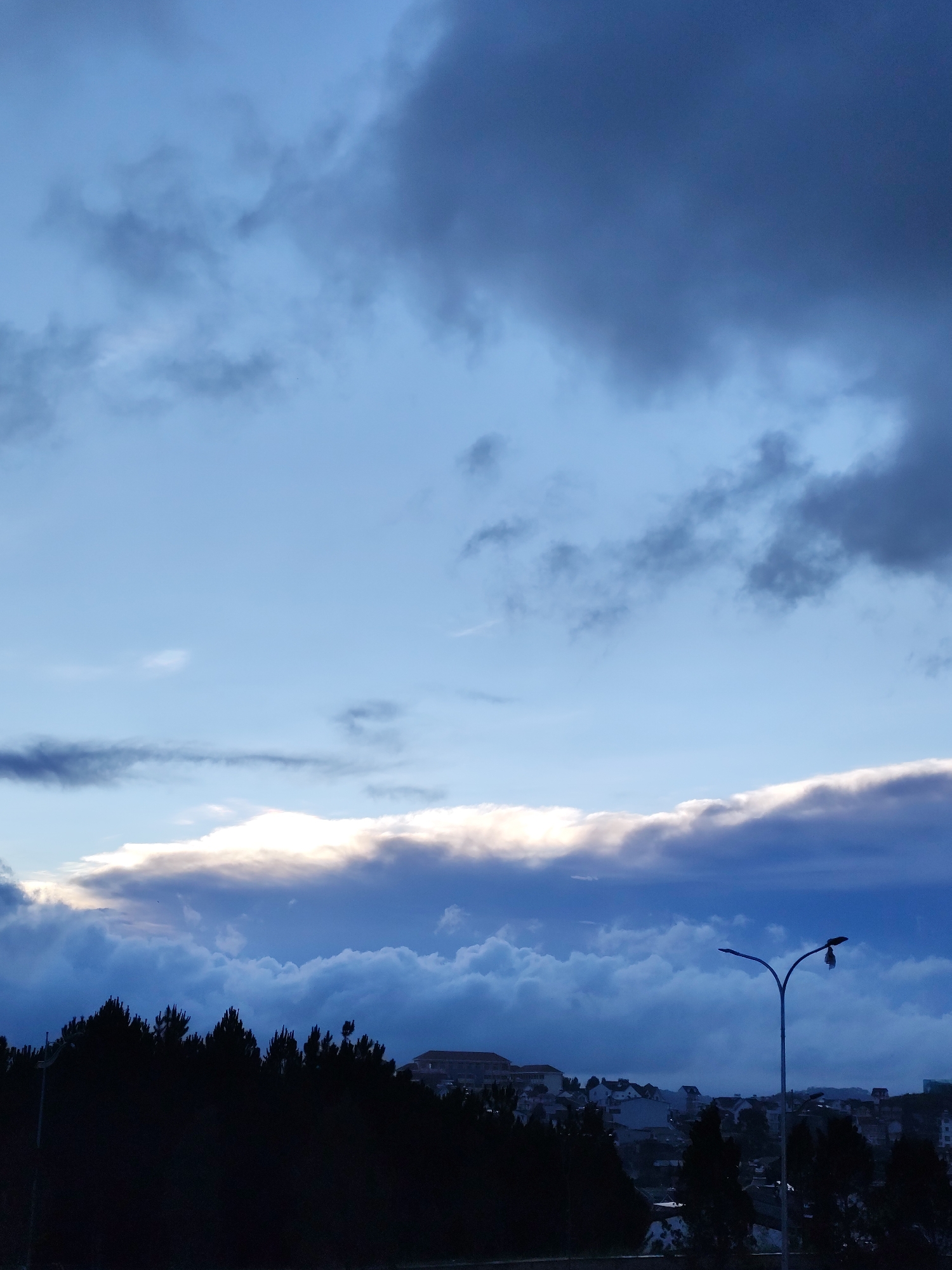 The image depicts a cityscape in Lang Biang - Da Lat, Vietnam, at dusk or dawn. The sky is a gradient of blues and grays, with a layer of clouds illuminated from behind, suggesting the sun is near the horizon. In the foreground, a dark silhouette of trees lines the horizon. A street light stands to the right, with a bent arm and a damaged lamp. Beyond the trees, a cluster of buildings, likely residential, is visible. The overall scene suggests a quiet moment at the edge of a city.