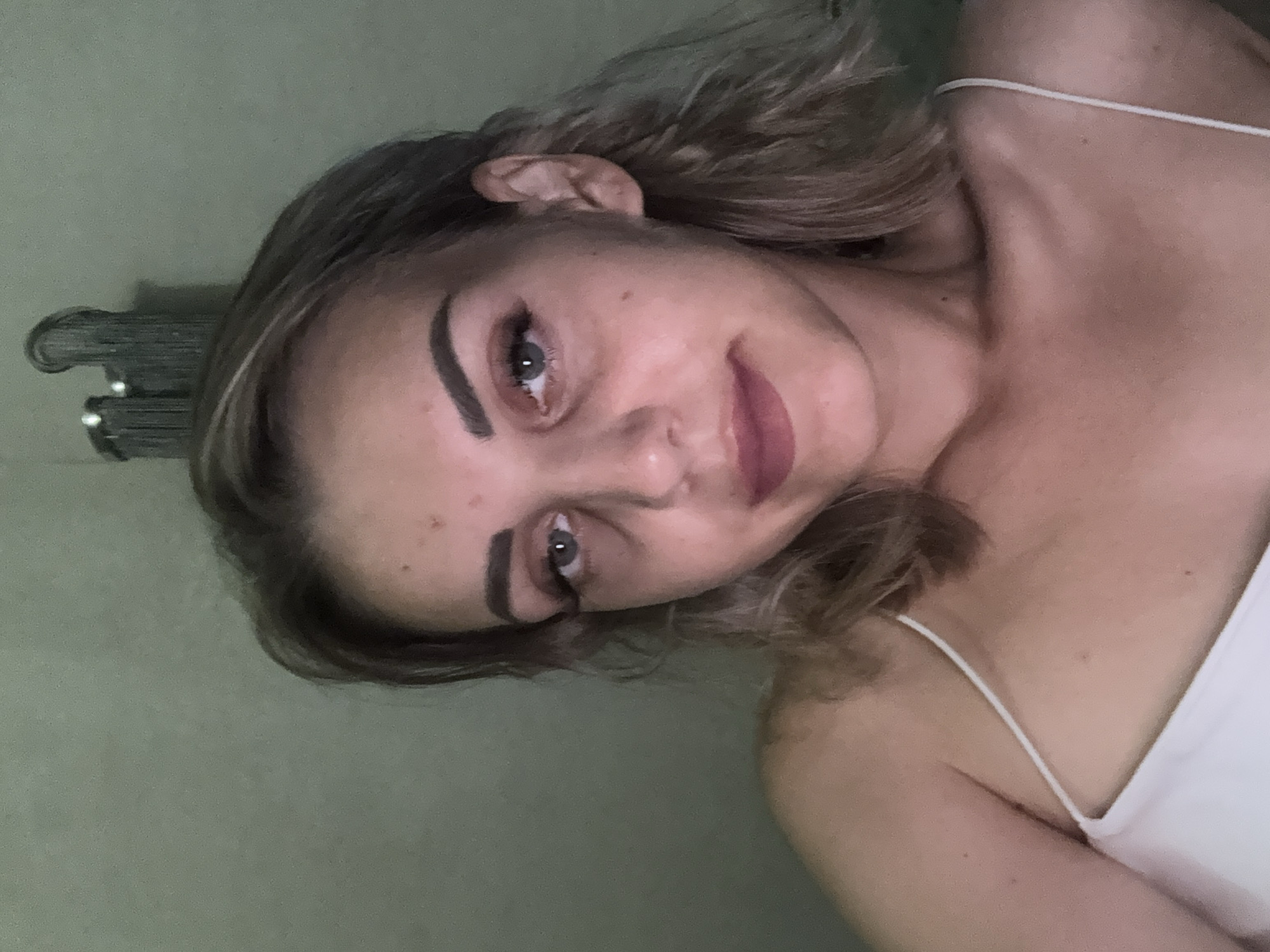 A young woman with blonde hair and fair skin is pictured in a close-up selfie. She is wearing a white spaghetti strap top. Her eyes are open and looking towards the camera, and her lips are slightly parted. The background appears to be a wall with a decorative light fixture visible in the upper right corner. There are no identifiable landmarks or elements that would suggest a specific location within the city of Yuzhno-Sakhalinsk, Russia.