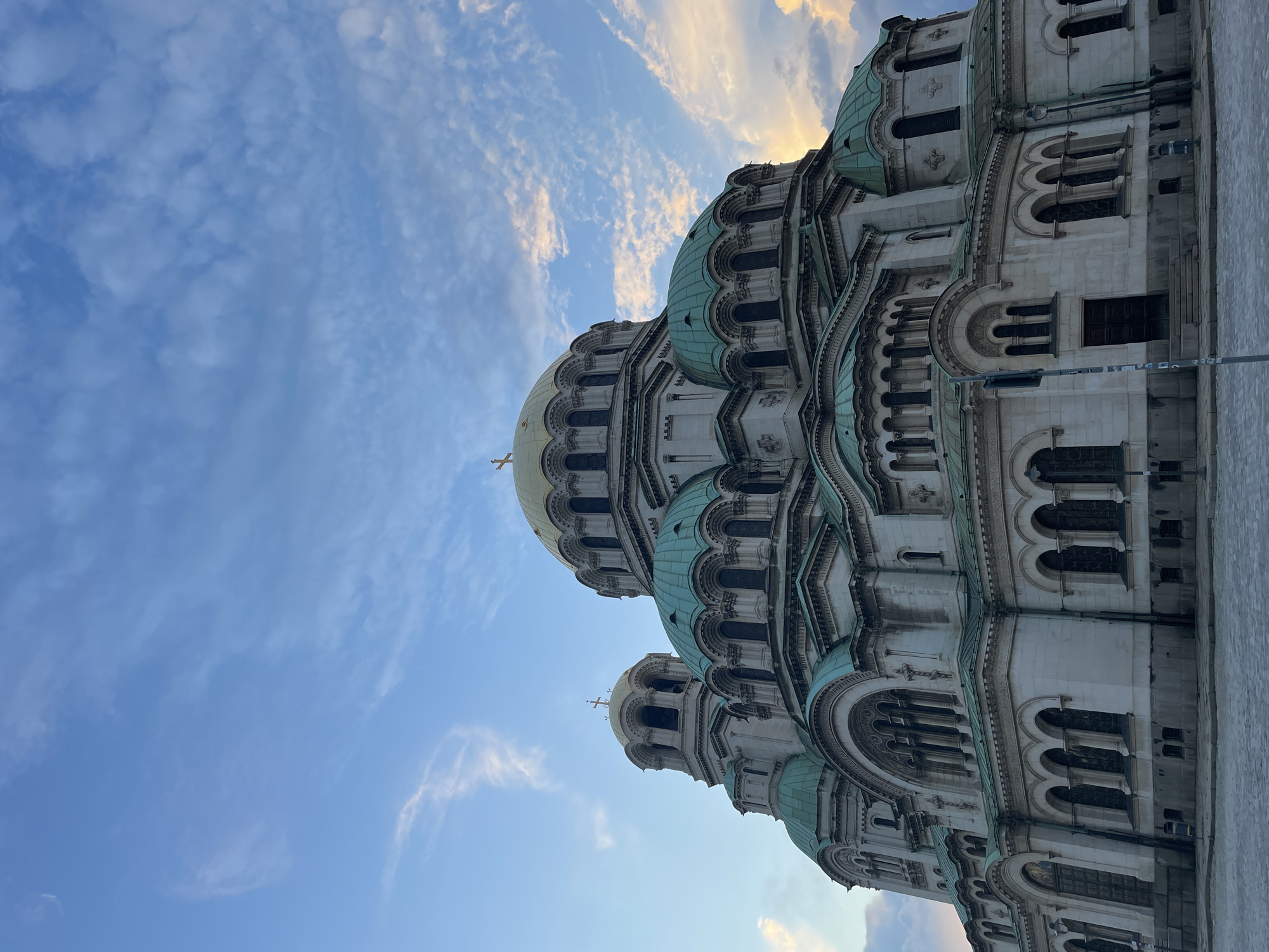 The image captures a low-angle view of the Alexander Nevsky Cathedral in Sofia, Bulgaria, against a dynamic sky. The grand Orthodox cathedral dominates the frame, showcasing its intricate architecture with light-colored stone facades, numerous arched windows, and decorative elements. Several domes are visible, with the central and prominent dome featuring a golden finish, while smaller domes exhibit a distinct greenish hue from patinated copper. Crosses adorn the peaks of the golden domes. The photograph emphasizes the scale and ornate detail of the building, with parts of its structure extending off the right edge of the frame.
The sky is a prominent element, displaying clear blue patches interspersed with scattered clouds. Some clouds are soft and white, while others, particularly towards the upper right, show hints of orange and pink, suggesting either a sunrise or sunset. The light seems to emanate from behind the cathedral on the right side, casting a soft glow on parts of the domes and highlighting their textures. No people or specific activities are visible within the frame. There is no visible text. The overall scene conveys the majestic presence of the cathedral under a picturesque sky during dawn or dusk.
