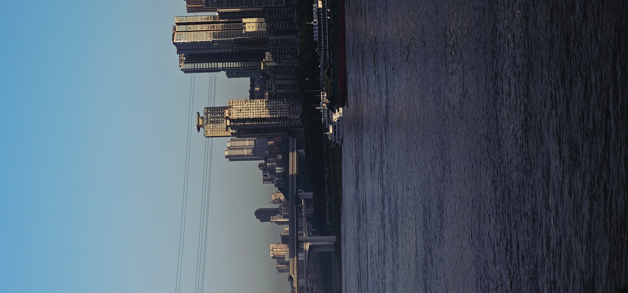 The image captures a waterfront cityscape in the Nan'an District of Chongqing, China. The foreground is dominated by a dark, rippling body of water, likely the Yangtze River. In the middle ground, a large cargo ship with a red hull is docked or navigating, accompanied by a smaller white passenger boat. Along the riverbank, a dense urban landscape rises, characterized by numerous high-rise buildings of varying architectural styles and heights. A prominent elevated highway or bridge structure spans across the scene in the mid-ground, supported by large concrete pillars, connecting different parts of the city. The sky is clear and a pale blue, indicating either early morning or late afternoon, with subtle power lines crisscrossing the upper portion of the frame. The lighting suggests the sun is low, casting soft shadows on the buildings. No individuals are clearly discernible. The overall impression is one of a bustling, modern metropolis situated along a major waterway.