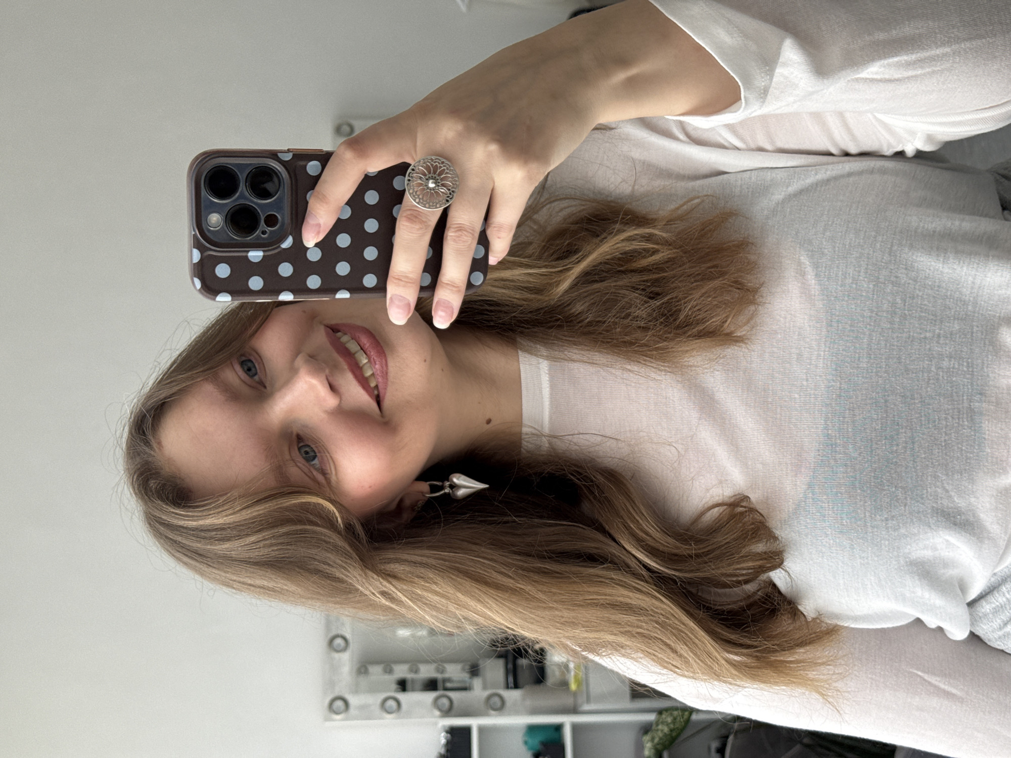 A young woman with light brown, wavy hair and blue eyes is captured in a mirror selfie, smiling broadly and cheerfully. Her head is tilted slightly to her right (viewer's left). She is wearing a white long-sleeved top, with a subtle light blue or gray undergarment visible beneath.
She holds a dark brown smartphone, likely an iPhone due to its triple-camera setup, in her right hand. The phone case features a light blue polka-dot pattern. On her right ring finger, she wears a prominent silver-toned ring with an ornate, circular filigree design. A silver-colored, heart-shaped earring dangles from her left ear. Her fingernails are neatly manicured with a clear or light pink polish.
The background, visible as a reflection in the mirror, shows a brightly lit indoor setting with white walls. Behind her, what appears to be a vanity table or shelf is reflected, displaying several small bottles and containers, along with a series of circular lights, suggesting a well-equipped dressing area or bathroom. The image portrays a casual and happy moment in an indoor environment, potentially in Izhevsk, Russia.
