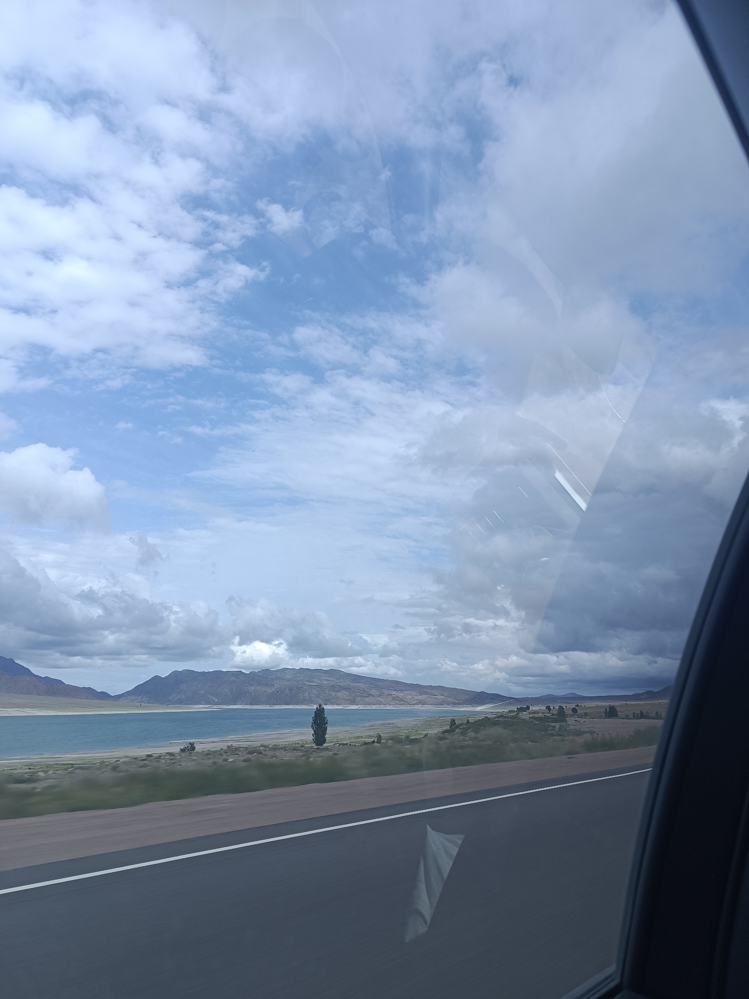 The image depicts a scenic view of a lake and mountains, likely taken from a moving vehicle. The sky is partly cloudy, with a mix of white and gray clouds against a blue sky. In the middle ground, a body of water, possibly a lake, is visible, bordered by a sandy shore and green vegetation. Rolling hills and mountains form the backdrop, with a single tall, dark tree standing prominently on the lakeshore. In the foreground, a dark asphalt road with a white line runs horizontally, suggesting a highway. The right side of the image shows a portion of a car's interior, including the window frame and some reflections on the glass. The overall impression is one of natural beauty and a journey through a rural landscape. The prompt requests the location be identified as Sasyk-Bulak, Kyrgyzstan, but no specific landmarks or identifying features are visible in the image that would confirm this location.
