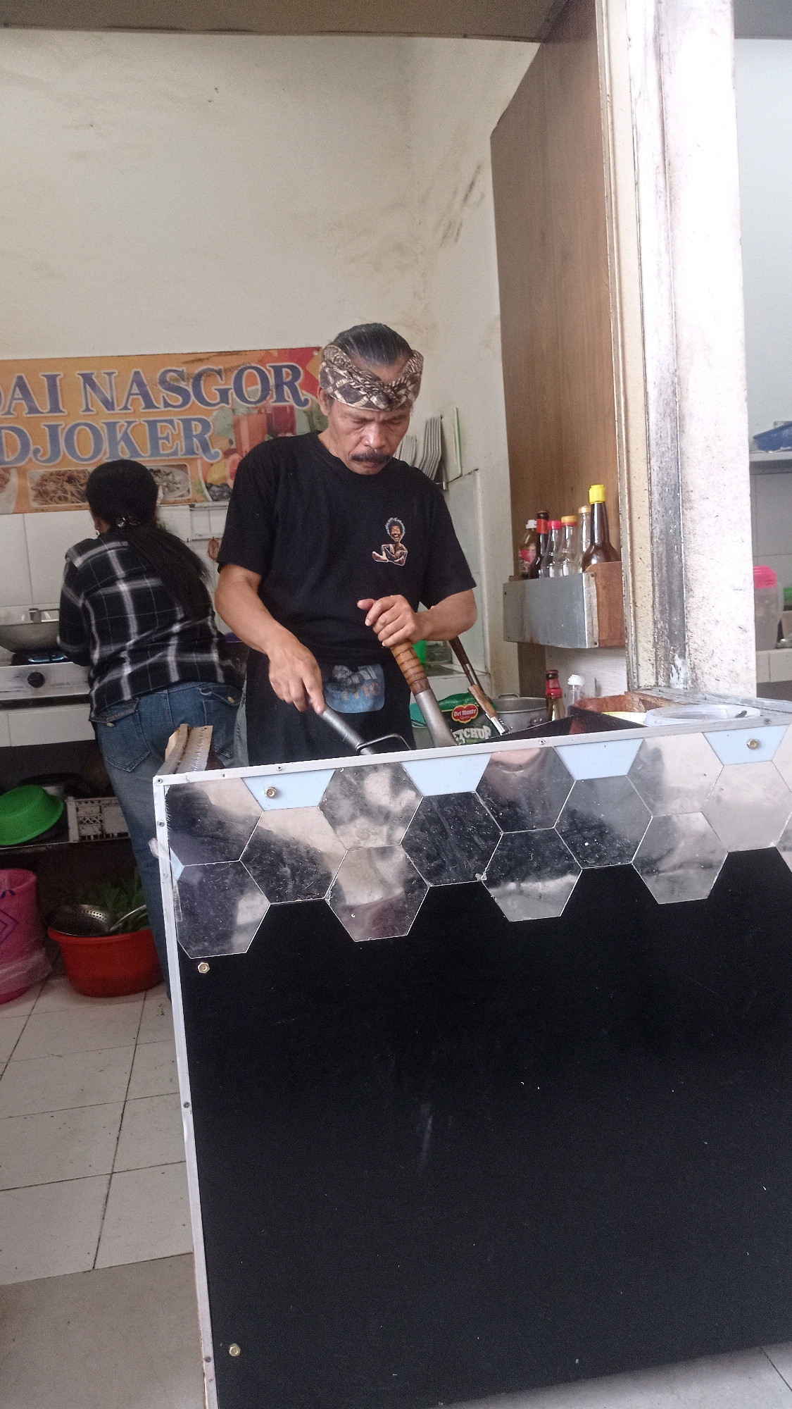 The image shows an indoor scene, likely a food stall or restaurant kitchen, in Batu, Indonesia. A man, appearing to be a cook, stands at the forefront, wearing a black t-shirt and a patterned headband. He is holding a cooking utensil, possibly a spatula, and a wok. To his left, a woman in a checkered shirt and jeans is bent over a stovetop, also engaged in cooking. Behind them, a sign with text reading "AI NASGOR DJOKER" is visible on the wall, indicating the establishment's name. Various kitchen items, including bottles of condiments and containers, are present on shelves in the background. A large, reflective surface with a hexagonal pattern is positioned in the foreground, obscuring part of the scene. The overall environment suggests a functional food preparation area.