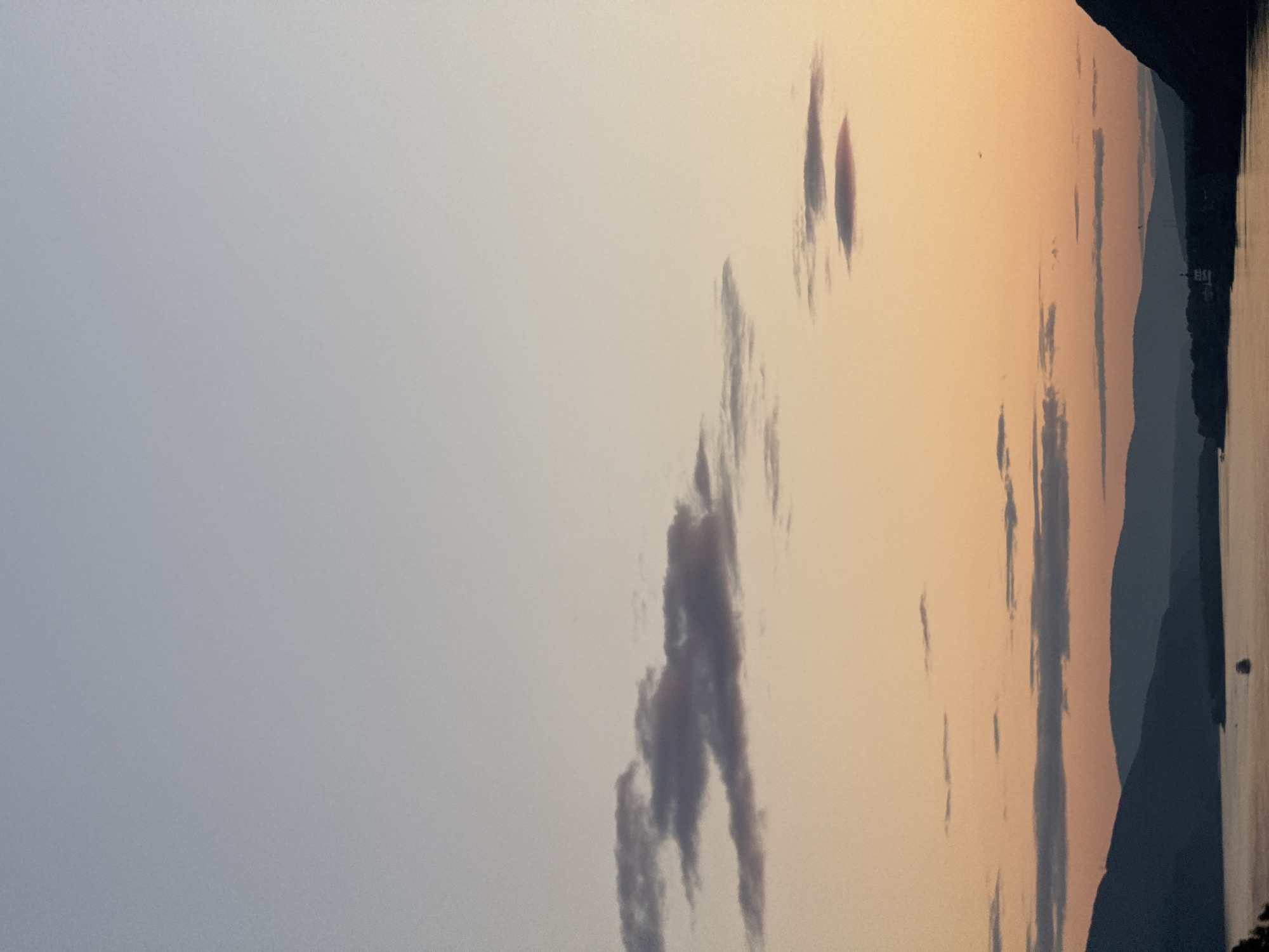 The image depicts a serene twilight scene over a body of water, likely a river, with silhouetted mountains in the distance. The sky transitions from a pale blue at the top to a warm orange and pink hue near the horizon, dotted with wispy clouds. In the foreground, dark mountains form a dramatic silhouette against the colorful sky. The water reflects the soft colors of the sky, and a small boat is visible on the water. A few buildings or structures can be discerned along the shoreline nestled amongst the mountains. The overall impression is one of tranquility and natural beauty at dusk. While the image is undated, the landscape is consistent with the scenery found in the vicinity of Krasnoyarsk, Russia, particularly along the Yenisei River, known for its picturesque mountain backdrops.