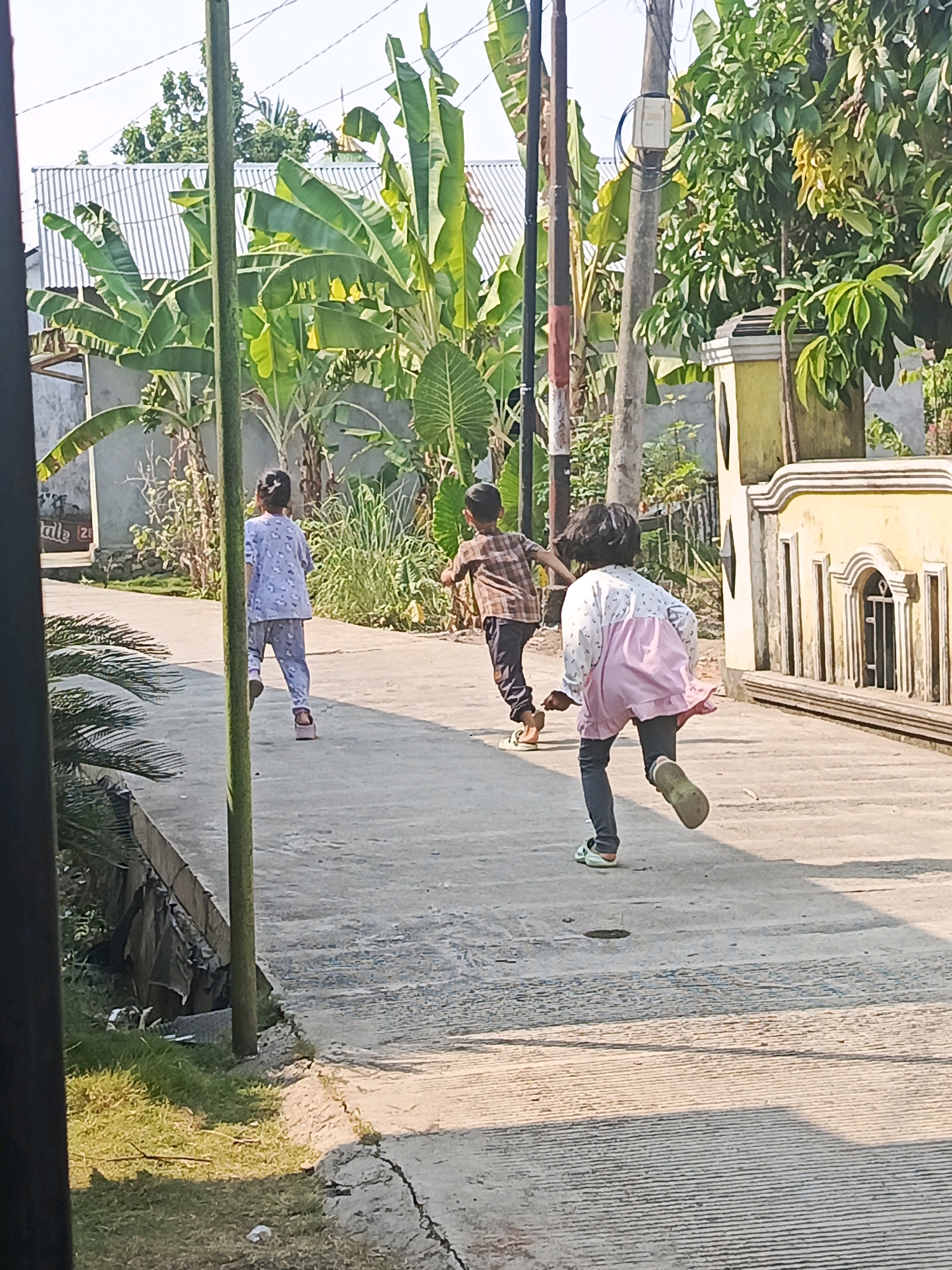 Three children are running on a paved path in what appears to be a residential area. The child in the foreground, facing away from the camera, wears dark leggings, a white top with a pink overlay, and light-colored sandals. The child in the middle, also facing away, is wearing a plaid shirt and dark pants. The child furthest to the left, facing away, is in light-colored pajamas. Behind the children, there is dense foliage, including banana trees, and residential structures with corrugated metal roofs. A decorative yellow wall with arched windows is visible on the right side of the image. The scene suggests a warm, sunny day. This image is likely taken in Lubuklinggau, Indonesia, based on common residential architecture and vegetation seen in the region.