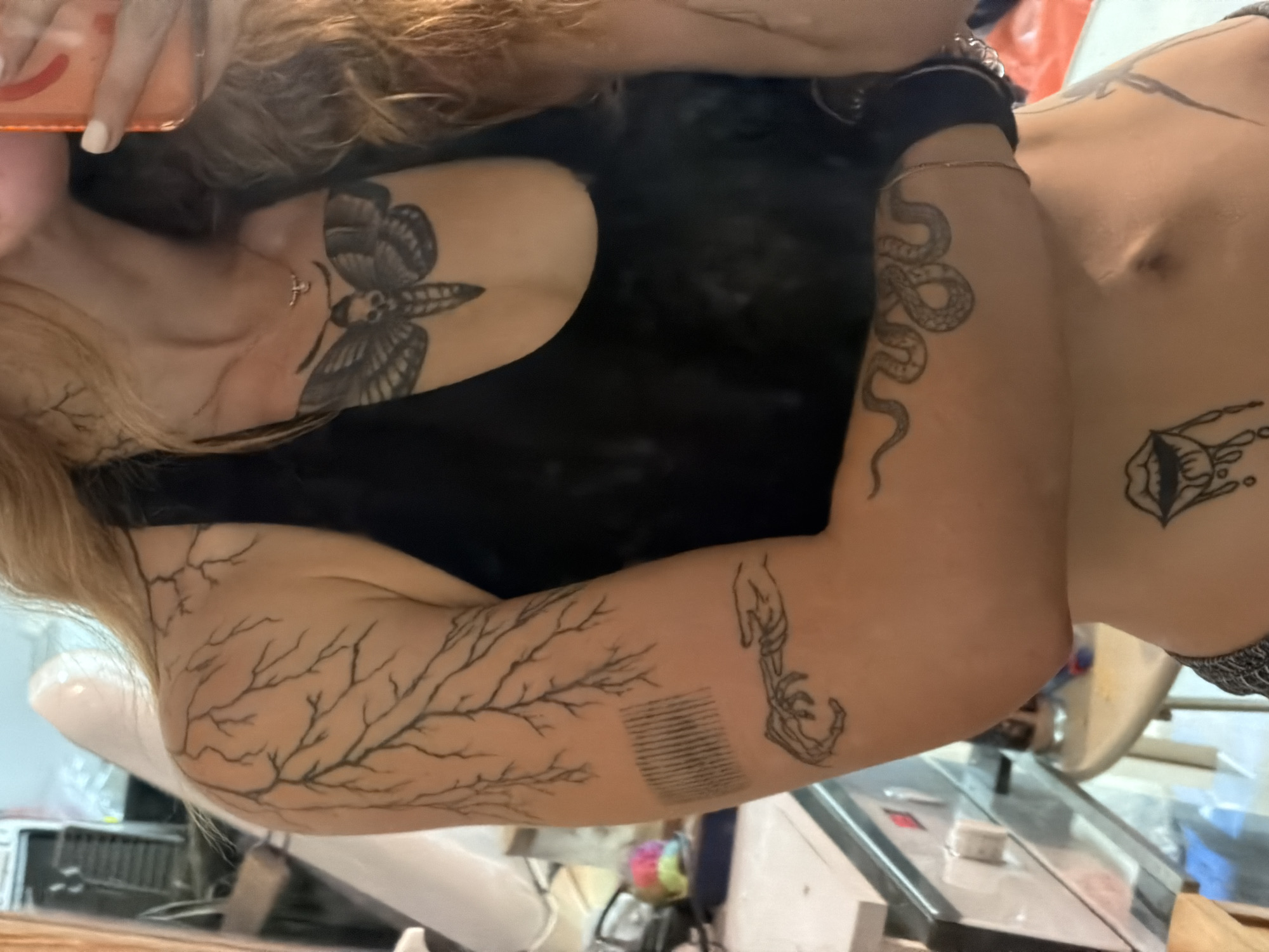 A close-up, medium shot captures a person from the chest down, predominantly focusing on their upper body and arm, adorned with multiple black ink tattoos. The subject, a woman with long, blonde, wavy hair, is wearing a black halter-neck top. Visible tattoos include a tree branch pattern extending across her shoulder and upper arm, a barcode-like design, and a skeletal hand reaching out on her forearm. A coiled snake tattoo wraps around her lower forearm, and a dripping mouth design is visible on her lower abdomen. On her chest, a large moth tattoo with a skull-like face is prominently displayed. The background appears to be an indoor environment, possibly a room with reflective surfaces, suggesting a mirror might be present, as implied by the partial reflection of the person and the surrounding details. The lighting is soft and diffused. There is no discernible indication of the location being Tel-Aviv, Israel.