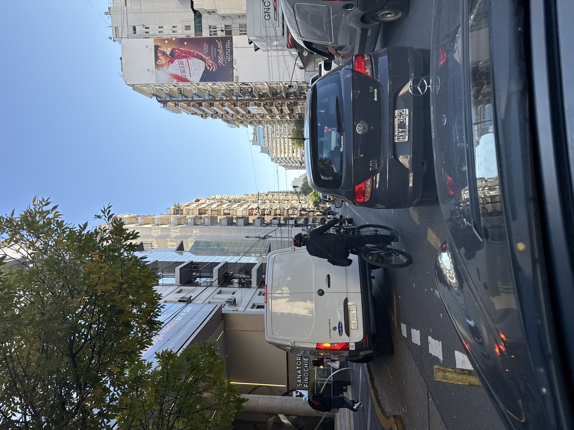 The image portrays a busy daytime street scene in a city, likely Buenos Aires, Argentina, given the visible cues. The viewpoint is from inside a vehicle, looking out onto a street filled with cars and buildings under a clear blue sky.
In the mid-ground, a dark Mercedes-Benz sedan with Argentinian license plate HXY 362 and "dietrich" branding is visible. Behind it is another dark sedan bearing "GOL", "AUTO ROD", and "Neumen" text. To the left, a white Mercedes-Benz Sprinter van with Argentinian license plate AH 735 VX is parked or stopped near a building entrance marked "SANATORI FINOCHIE", indicating Sanatorio Finochietto. A person on a bicycle, wearing a helmet, is cycling past the van, and another pedestrian is seen near the sanatorium entrance. A gray van with "GNC" on its side is also present in the traffic. Reflections of the car's interior are visible on the right side of the image.
The street is flanked by multi-story buildings, some with balconies. One prominent building features a large billboard advertising "Michael 23 DE ABRIL SOLO EN CINES". Another building also displays "GNC" text. Green trees with dense foliage are in the foreground on the left. The road has white painted square markings and a yellow curb line. The overall scene depicts typical urban traffic and activity on a clear, sunny day.