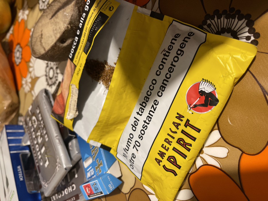 The image captures a close-up, top-down view of several items resting on a patterned surface. The main subject is an open yellow pouch of "AMERICAN SPIRIT" loose-leaf tobacco, spilling some of its contents. The pouch features a logo of a person with a feathered headdress smoking a pipe.
Visible text on the tobacco pouch, in Italian, includes the health warning: "Il fumo del tabacco contiene oltre 70 sostanze cancerogene" (Tobacco smoke contains over 70 carcinogenic substances) and another warning related to "bocca e alla gola" (mouth and throat). A helpline number "verde 800.554.088 per smettere di fumare" (toll-free 800.554.088 to quit smoking) is also displayed. The pouch indicates a weight of "30g e" and the product type "CIGARETTE TOBACCO".
Other objects in the frame include a silver-wrapped bar of chocolate with "CHOCOLAT" clearly visible, and a small blue box of "SLIM" "120 FILTER TIPS" with text "PLASTICA DEL FILTRO" (plastic filter). A dark item labeled "WISTECH" and a wooden object are partially visible in the background. The items are arranged on a brown surface adorned with large orange, white, and dark floral or geometric patterns, possibly a tablecloth or placemat. The scene appears to be indoors, likely in Dosso (Ciappanico), Italy, based on the provided context and Italian text.