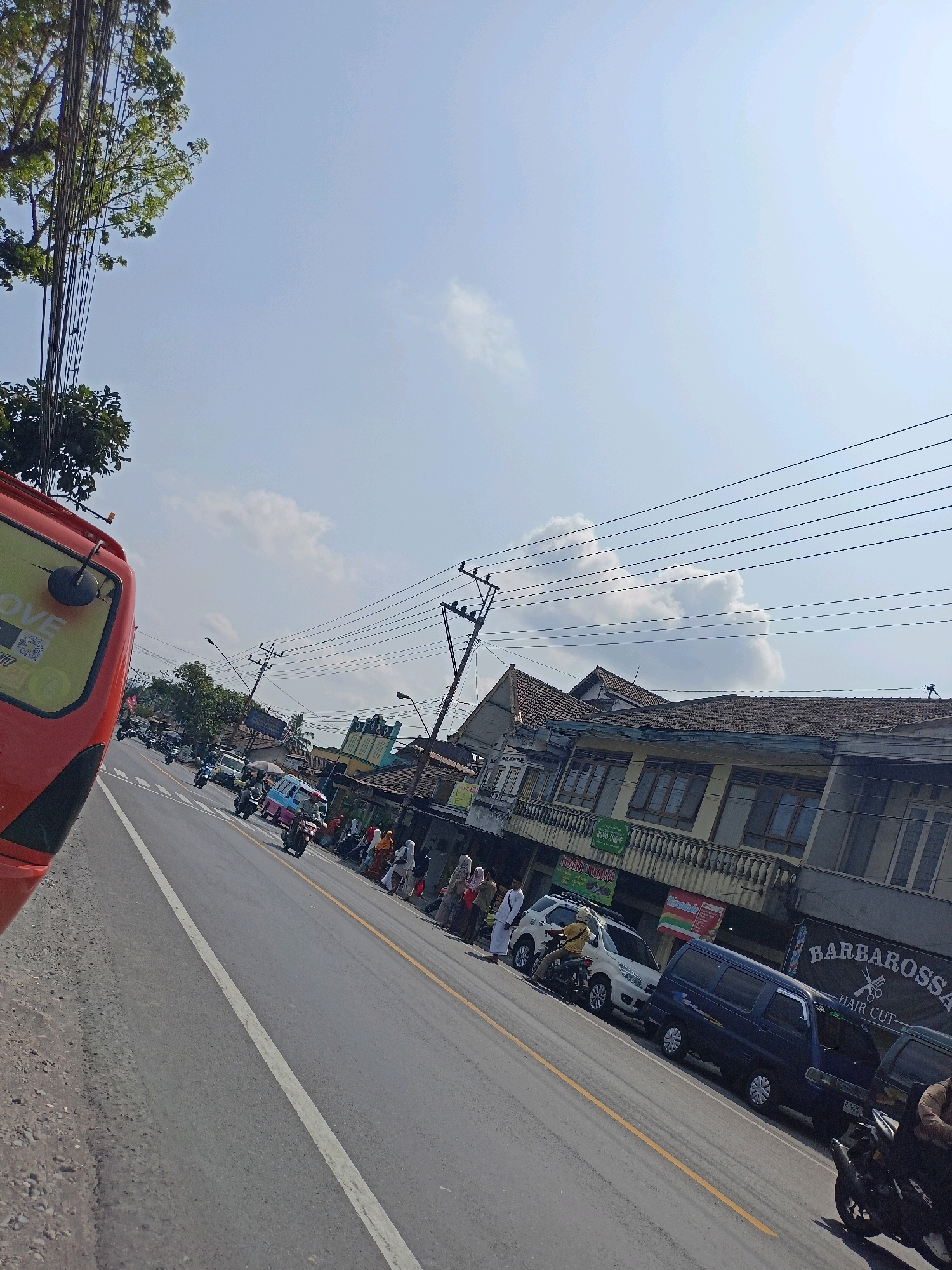 The image depicts a street scene in Payaman, Indonesia, on a bright, clear day with scattered clouds. A red vehicle, partially visible on the left, is positioned close to the edge of the road. The road itself is paved and marked with a white center line and a yellow lane line.
Along the right side of the road, a row of buildings with tiled roofs and balconies lines the street. Several people, some wearing headscarves, are seen standing or walking on the sidewalk in front of these establishments. Various vehicles, including cars, scooters, and a small bus, are parked or moving along the road. Signs for businesses, such as "BARBAROSSA HAIR CUT," are visible on the buildings. Overhead, a network of electrical wires stretches across the sky, supported by utility poles. The presence of trees with green foliage is also noted.