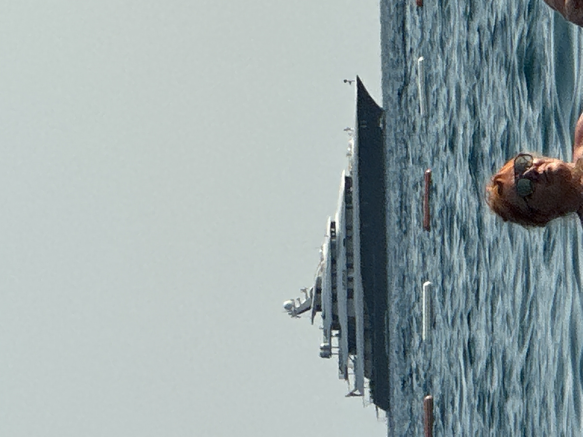 A luxury yacht is visible on the horizon, with a pale blue sky in the background. In the foreground, the heads and shoulders of two people are visible, partially submerged in the blue water. One person is wearing sunglasses. There are several buoys floating in the water. The scene appears to be a coastal area.