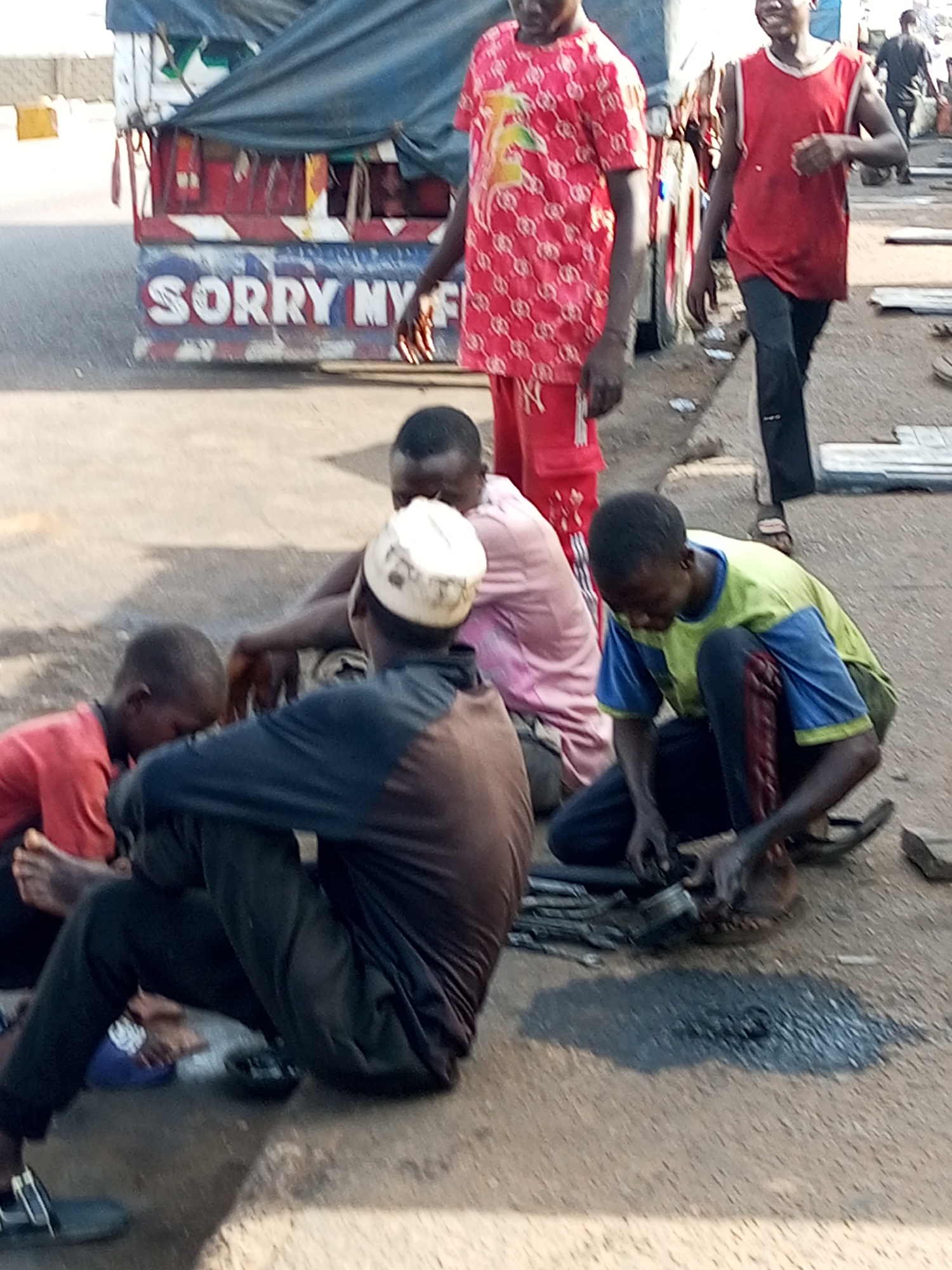 The image depicts a group of young men gathered on a dusty, unpaved ground in the city of Gombe, Nigeria. In the background, a truck is partially visible, with the words "SORRY MY F" painted on its side. Several of the men are seated or crouched on the ground, seemingly engaged in work or conversation. One individual, wearing a red shirt with a white pattern, stands and walks away from the viewer. Another person, in a red tank top and dark pants, walks towards the right of the frame. Two of the seated figures appear to be working on something on the ground, possibly a mechanical object. The overall atmosphere suggests an outdoor, informal gathering or work setting, likely in a commercial or industrial area of Gombe.