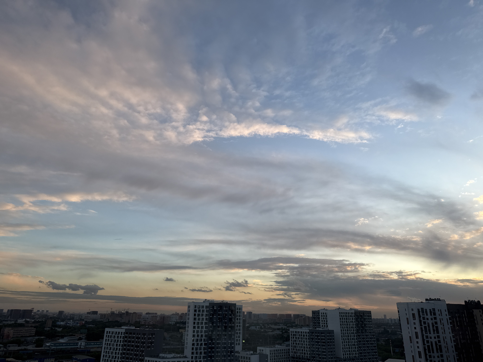 This is a wide-angle, eye-level photograph of a cityscape at dusk, with a dramatic sky filled with clouds. The foreground is dominated by several modern, multi-story apartment buildings with white and gray facades. These buildings are clustered together, forming a dense urban landscape that extends towards the horizon. In the background, more buildings of varying heights are visible, some appearing to be under construction with cranes dotting the skyline.
The sky is the most striking element of the image. It is a canvas of blended colors, transitioning from a pale blue at the top to soft oranges, pinks, and grays as it nears the horizon. Wispy, elongated clouds stretch across the sky, catching the fading sunlight and creating a textured, layered effect. Some clouds are tinged with a golden hue, suggesting the last moments of sunset, while others are darker, hinting at approaching evening or lingering weather patterns. The overall mood is serene and slightly melancholic, capturing the transition from day to night over a developing urban area. No people are visible in the image. The location context suggests this is a view of residential buildings in Бордуки, Russia.