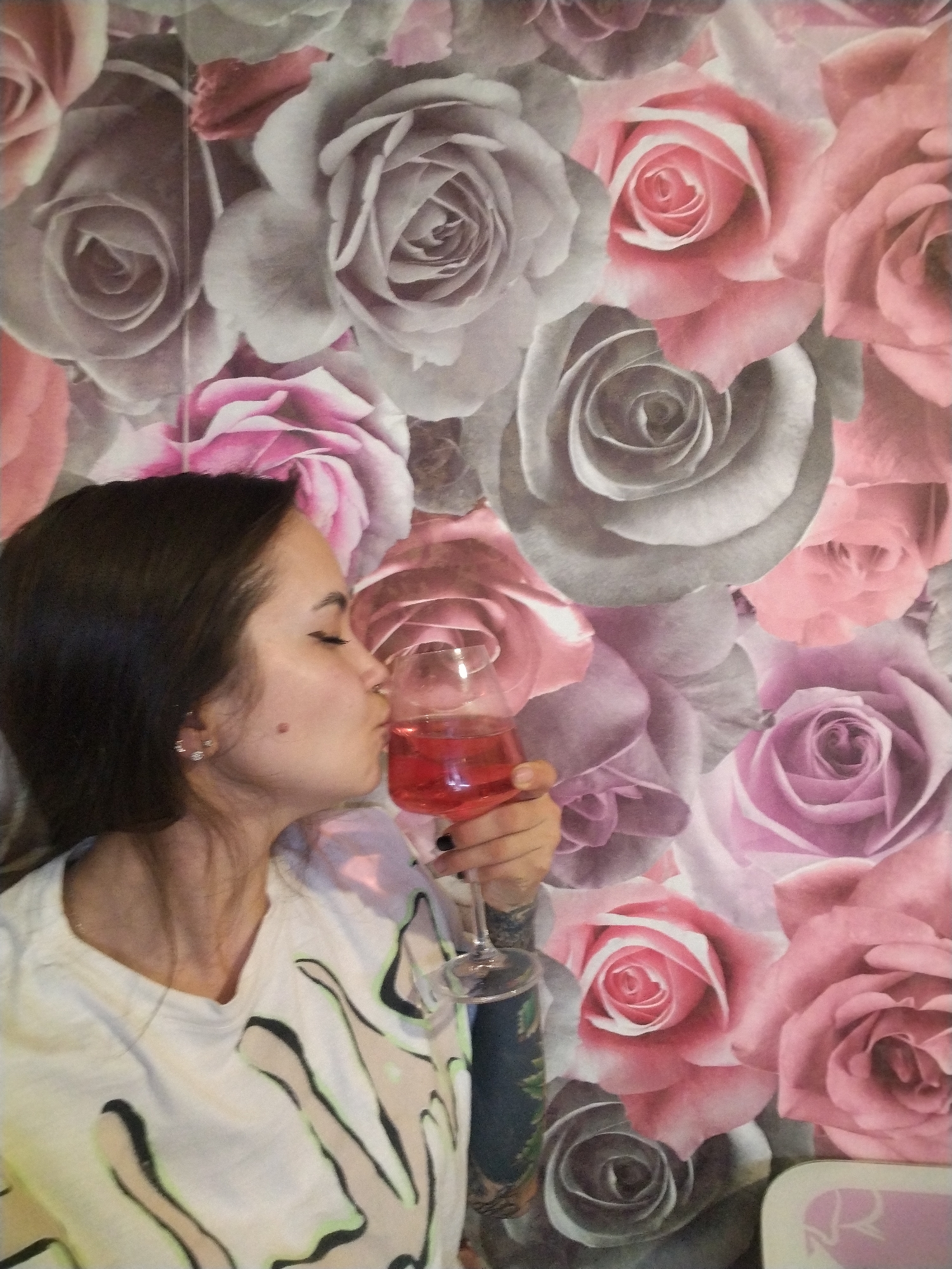 A woman with dark hair is holding a glass of red liquid to her lips in front of a wall covered in a pattern of pink, grey, and purple roses. She wears a white t-shirt with a stylized drawing. Her arm with a tattoo is partially visible. The scene suggests an indoor setting. No information suggests that the location is in the city of Noviy, Russia.