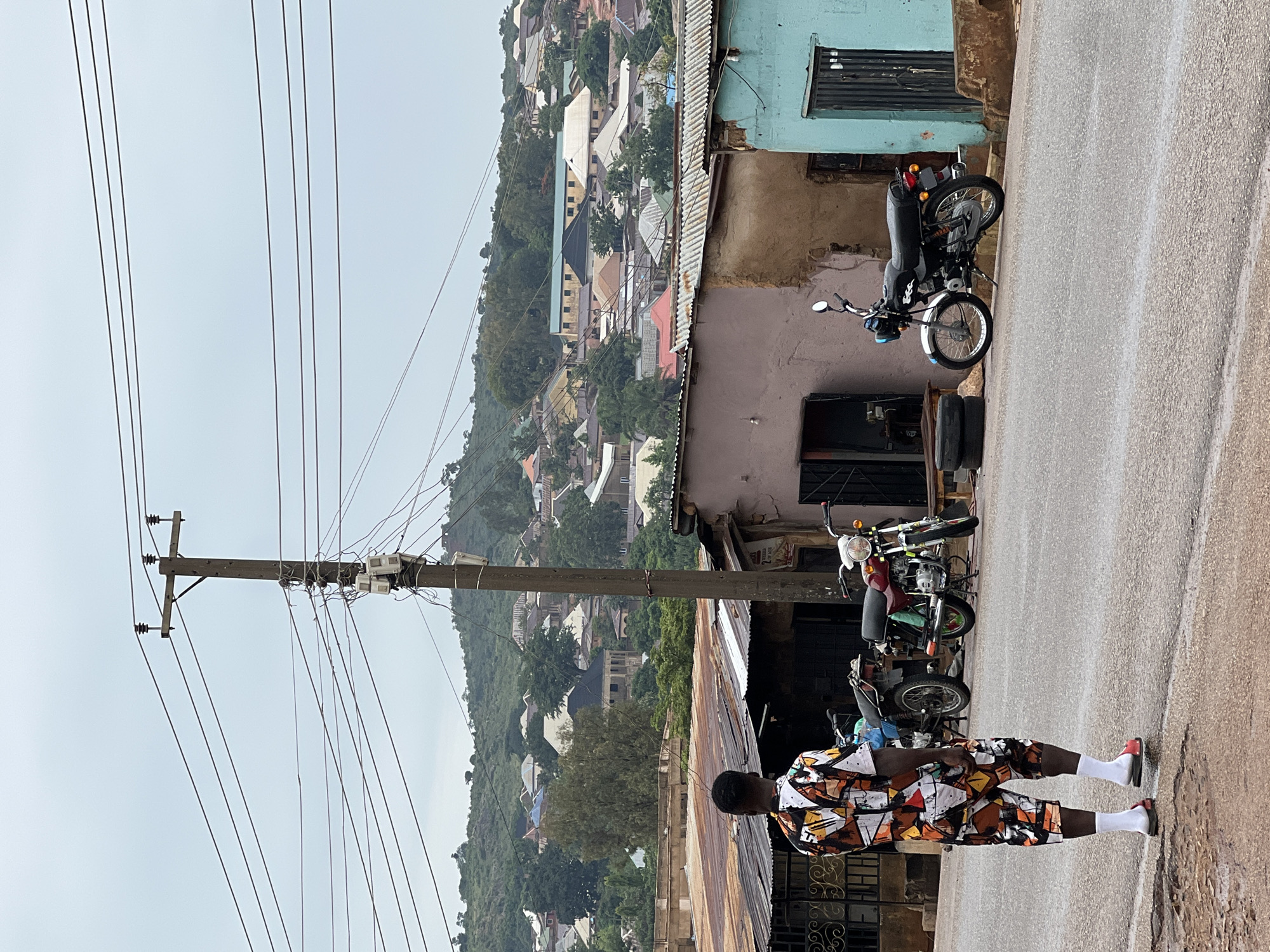 The image depicts a street scene in Jos, Nigeria. An individual wearing a patterned shirt and shorts, white socks, and sandals stands on a paved road with a telephone pole and associated wires to the left. Several parked motorcycles are visible along the roadside. In the background, a hillside is covered with densely packed buildings and vegetation. A variety of building materials and designs are visible in the background, and the sky is overcast.