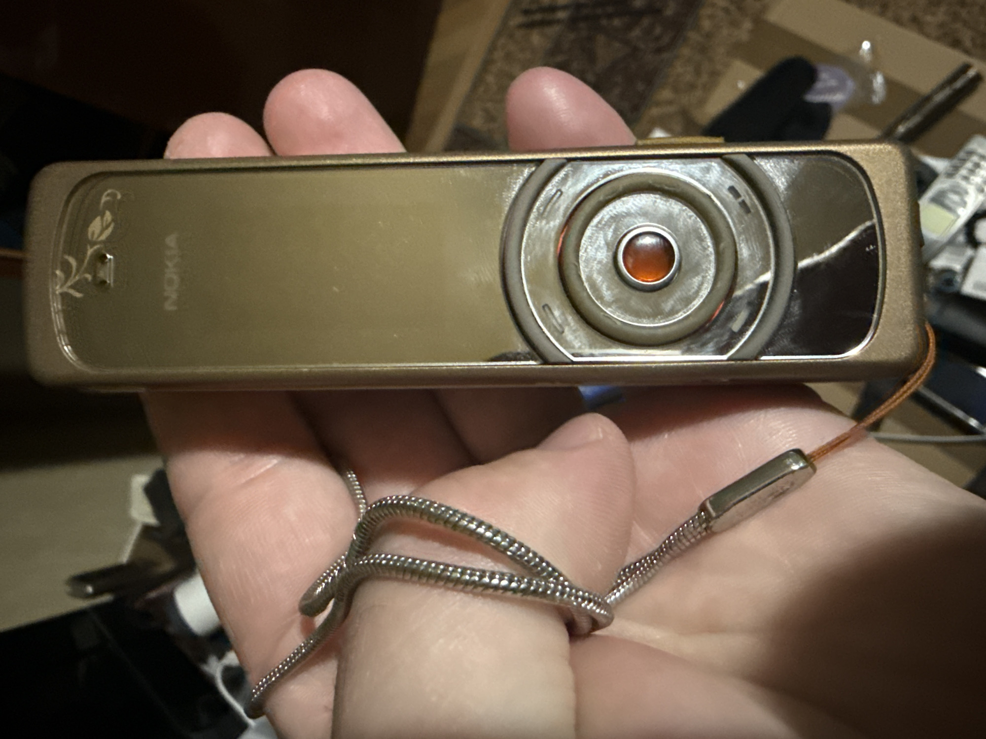 The image displays a person's hand holding a golden-bronze Nokia mobile phone horizontally. The phone features a decorative etched floral design on its left side and the "NOKIA" brand name. The right side is dominated by a circular camera module, distinguished by a prominent amber-colored jewel at its center, surrounded by concentric metallic rings. A silver-colored, braided metallic wrist strap or lanyard is looped around the fingers, connecting to the phone via a thin brown cord. The background is blurred, suggesting an indoor environment, possibly a room with a patterned floor or carpet. Faint glimpses of other electronic devices, including another mobile phone, are visible in the background. The image focuses solely on the phone and the hand holding it; no specific activity beyond holding the device is depicted, nor are any emotions discernible. The visible text is "NOKIA". The location context is Krumovgrad, Bulgaria.