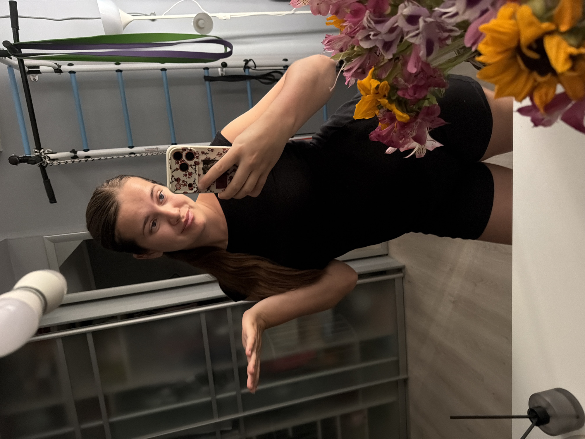 A young woman with long, brown hair pulled back in a ponytail, wearing a black t-shirt and black shorts, takes a selfie in a mirror. She is standing in a room with a wooden floor and a large, mirrored closet. To her right, there is a wall-mounted pull-up bar with blue bars and resistance bands hanging from it, along with a floor lamp. In the foreground, a bouquet of flowers, including pink alstroemeria and a sunflower, is visible on a white surface, likely a vanity or table. The woman is holding a smartphone with a floral case in her right hand, and her left hand is extended outwards with her palm up. The overall impression is that of a domestic setting, possibly a home gym or a bedroom, within the city of Sochi, Russia.