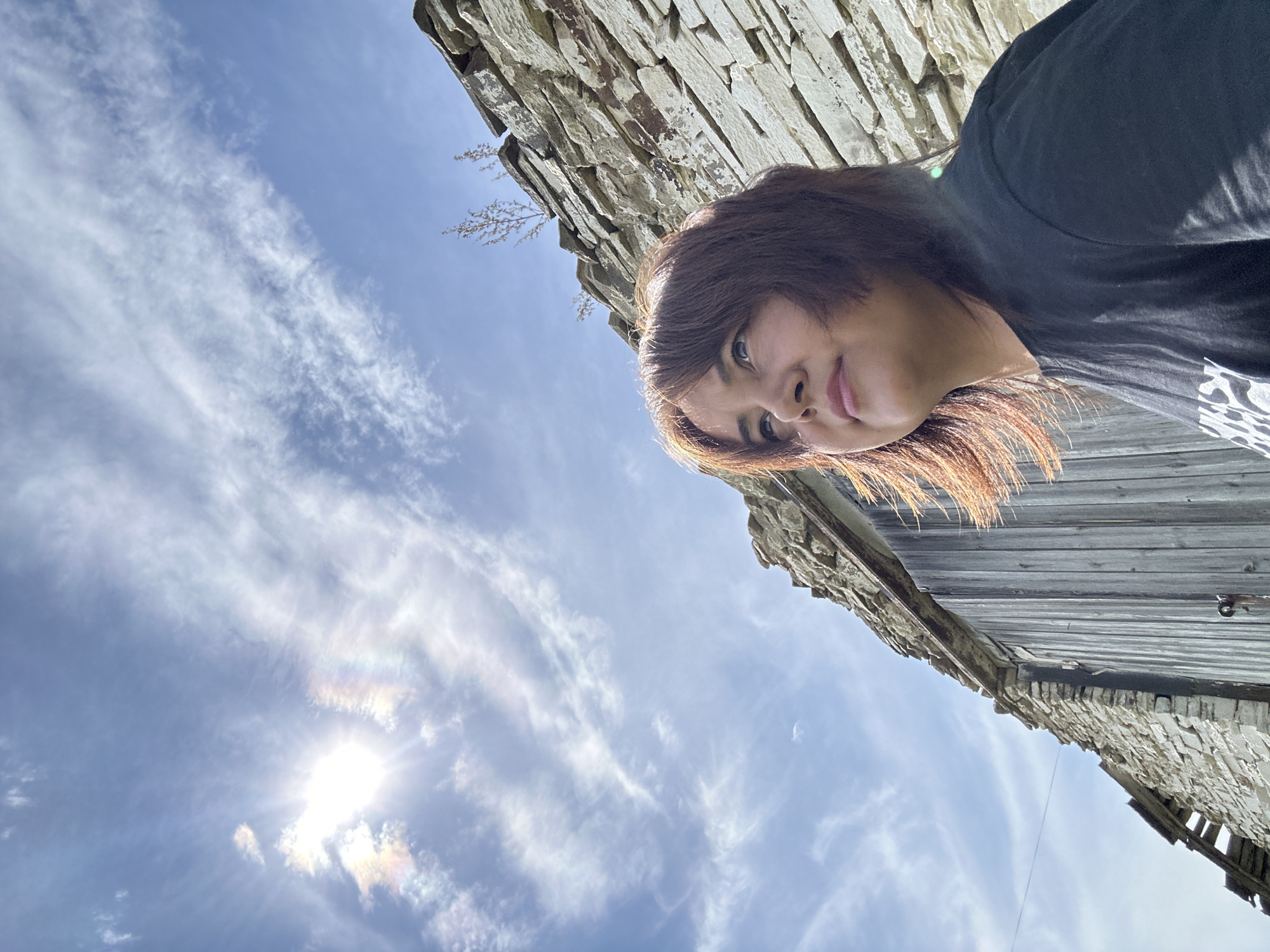 A medium shot shows a woman with reddish-brown hair looking up and to the right, positioned in the lower right of the frame. She is wearing a black t-shirt. Behind her, a weathered stone wall and wooden structure are visible, suggesting an old building. The sky above is blue with wispy clouds, and the sun is bright and partially obscured by clouds, creating a rainbow-like halo effect. The scene appears to be outdoors.