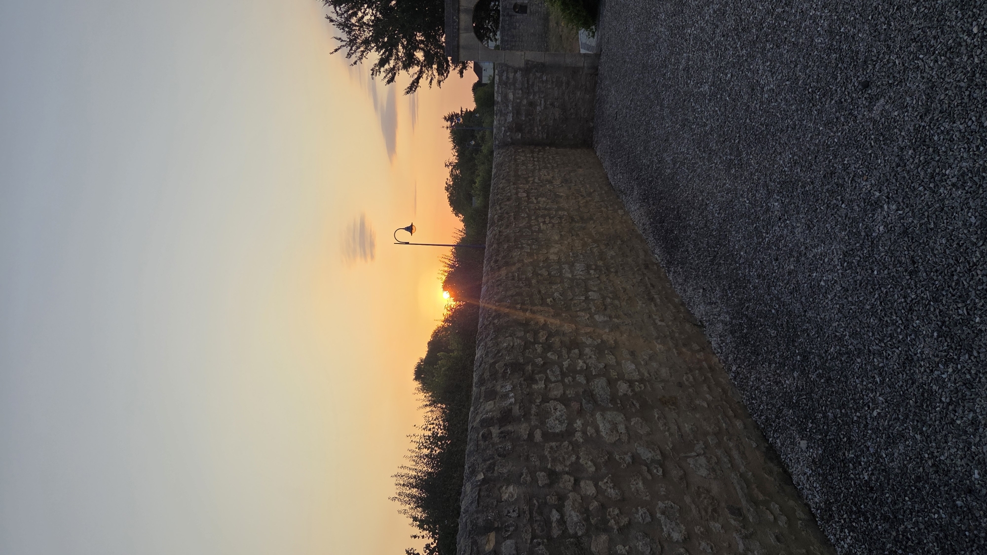 The image depicts a sunset scene in the city of Macé, France. A stone wall runs diagonally across the foreground, separating a gravel-covered area on the right from a grassy area with trees on the left. The sun is setting behind a line of trees and a street lamp is visible to the right of the sun. The sky is a gradient of orange and yellow hues, with a few wispy clouds. The overall impression is one of a peaceful, rural landscape at dusk.