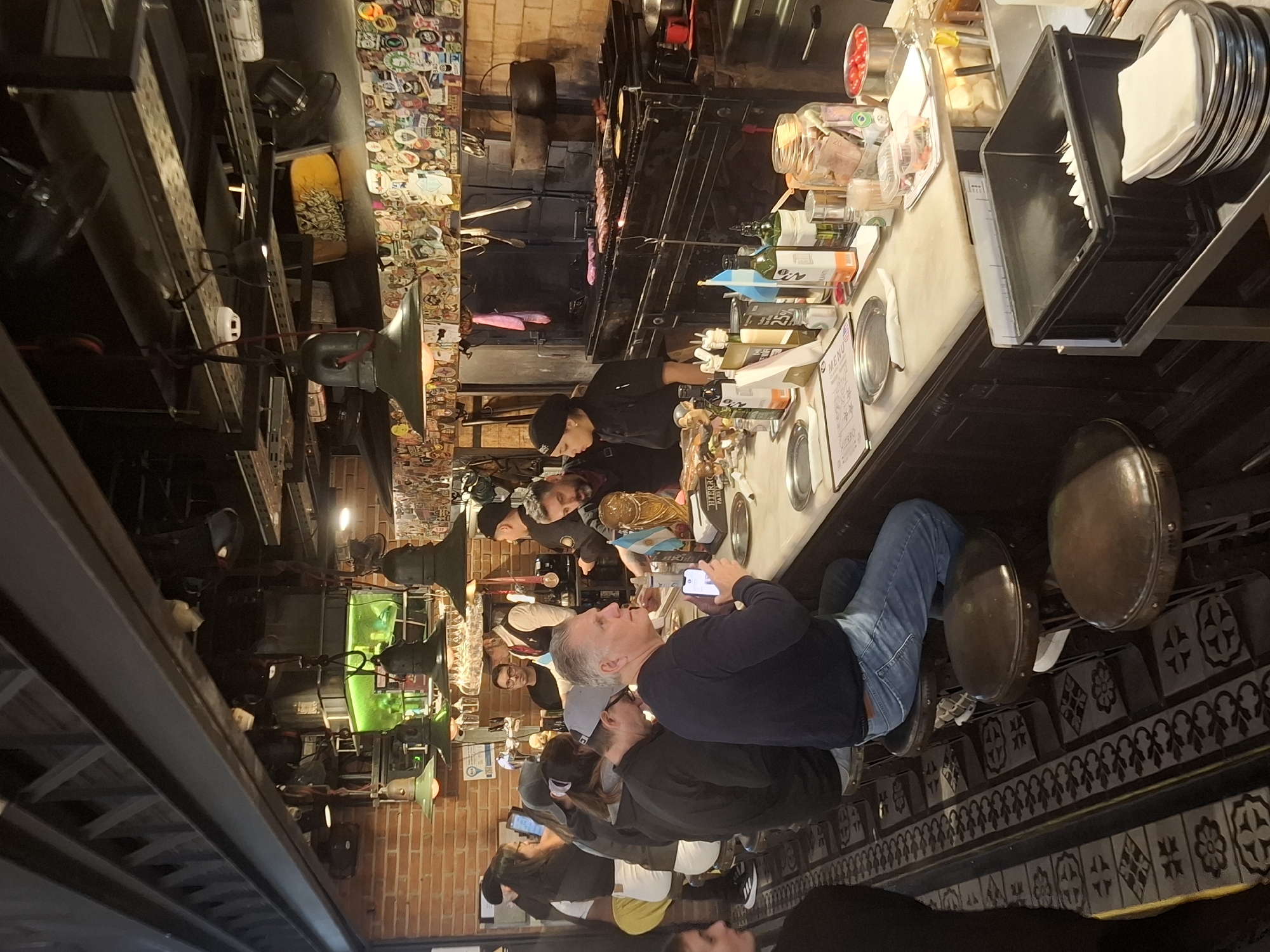 The image depicts a busy scene inside a restaurant. Several people are seated at a counter, with some looking at their phones. The counter has tableware, including plates and serving platters. Behind the counter, restaurant staff are visible preparing food, possibly grilling meat over an open fire. The interior of the restaurant is decorated with exposed brick walls and a variety of lighting fixtures. There are also decorations on the walls, including stickers and other memorabilia. The setting is likely a restaurant in Buenos Aires, Argentina.