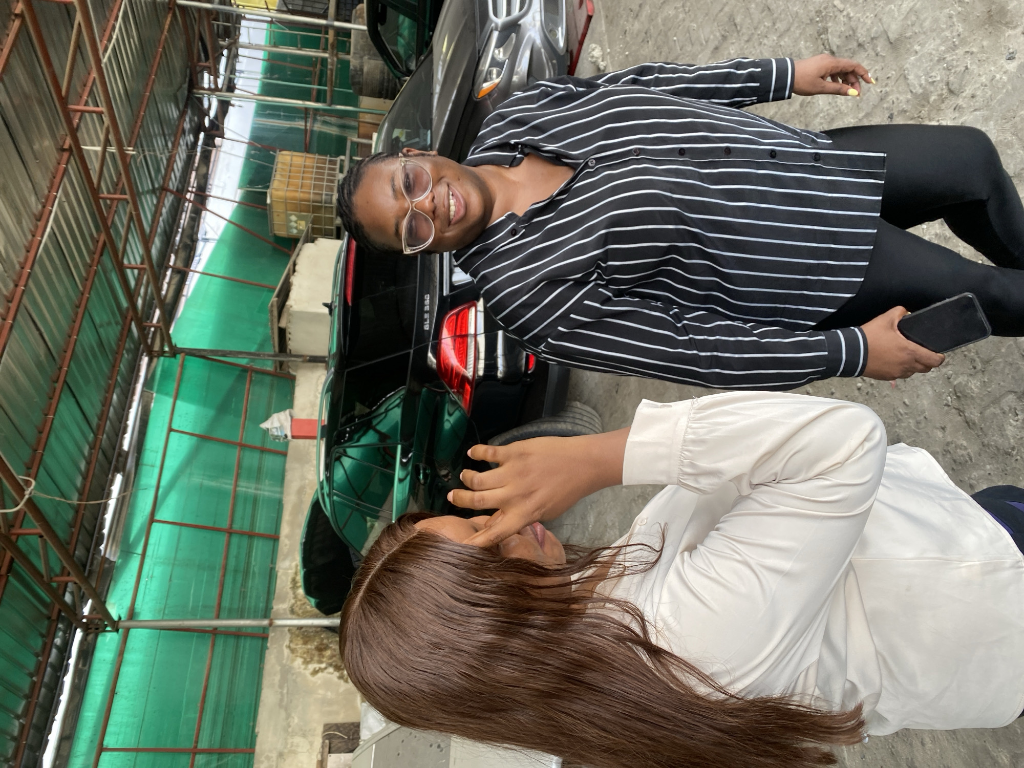 In Igboefon, Nigeria, the scene captures two women outdoors, next to two cars. One woman, wearing glasses and a black-and-white striped shirt with black pants, stands smiling and holding a phone. Behind her is a dark-colored SUV, possibly a Mercedes-Benz GLE 350, parked near a green-covered structure with metal supports. The second woman, wearing a light-colored blouse, is lying down with her hand touching her face. She has long brown hair and is close to a dark green car. The ground appears to be concrete or gravel.