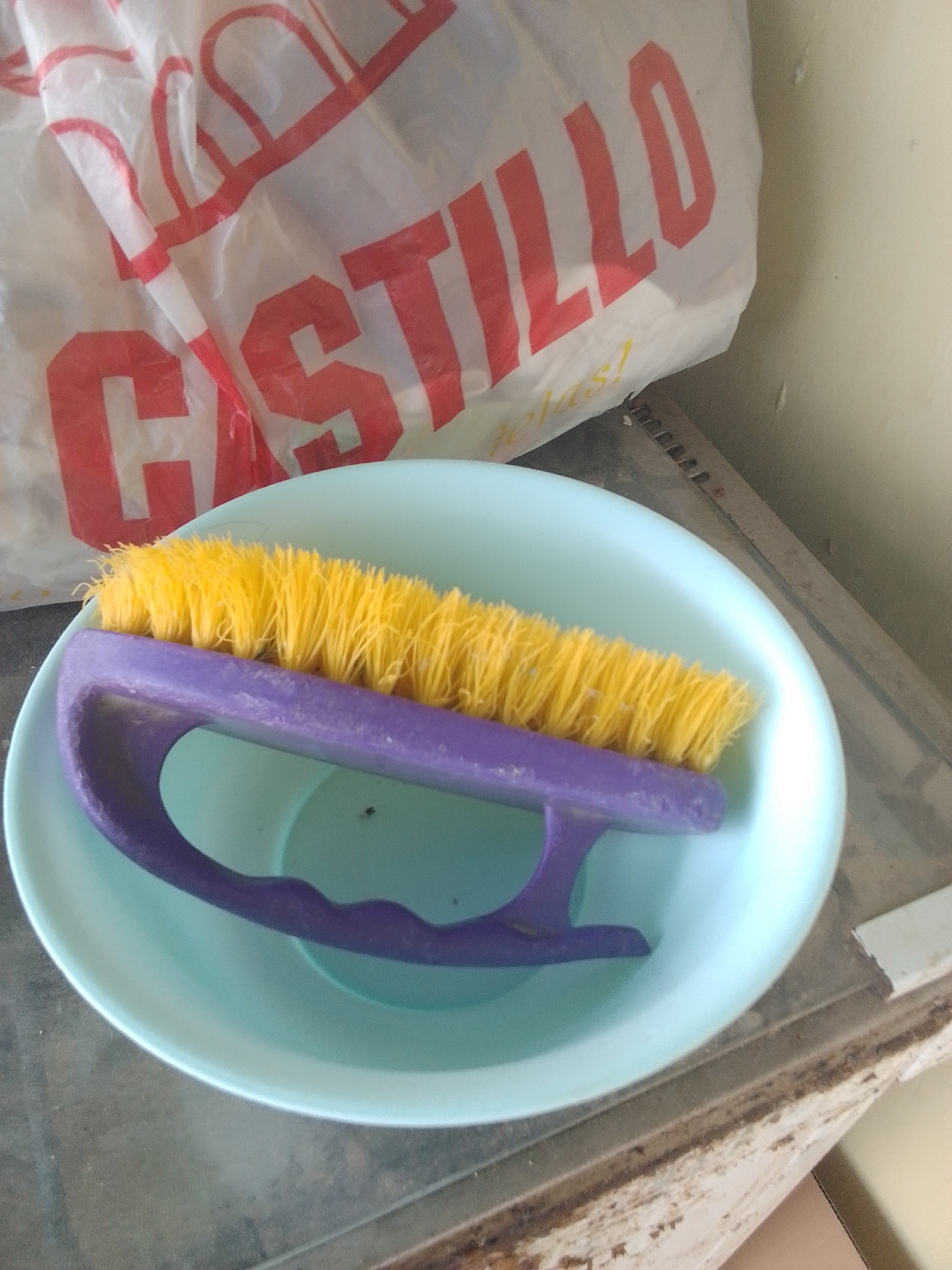 A yellow-bristled scrub brush with a purple handle rests in a pale blue bowl. The bowl and brush are positioned on a surface that appears to be a shelf or countertop, which has some visible wear and discoloration. In the background, a white plastic bag with large red lettering is draped behind the bowl. The visible text on the bag reads "CASTILLO". The lighting suggests an indoor setting, possibly during the daytime, with a neutral background wall.