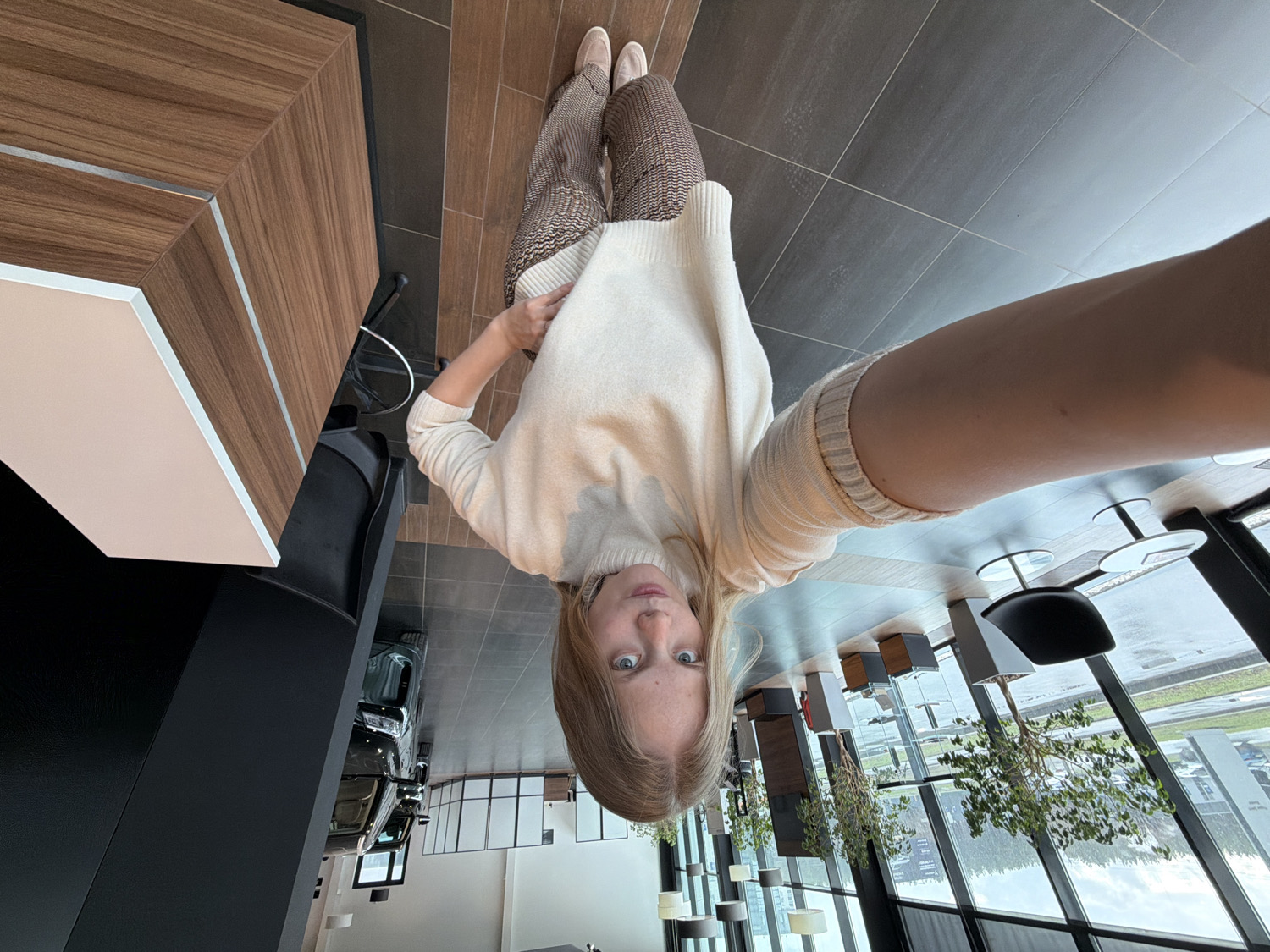 A high-angle, slightly tilted selfie-style shot captures a young woman looking directly at the camera in a modern indoor setting. The woman, with light brown hair parted down the middle and blue eyes, is wearing a cream-colored turtleneck sweater, patterned brown and white trousers, and light-colored flat shoes. Her left hand is casually placed near her waist, holding her sweater, while her right arm extends out, likely holding the camera.
The indoor space features contemporary design with dark grey tiled flooring and a prominent wooden-patterned counter or bar area with a light cream top edge. A black, sleek pillar or structure is visible to the left. In the background, large floor-to-ceiling windows reveal an overcast or rainy outdoor scene. Through the windows, blurred trees, a wet ground, and what appears to be a car showroom or dealership are visible, with at least one dark-colored car (possibly an SUV or jeep) parked inside, near the glass. There are also tables with black chairs or stools scattered throughout the spacious interior. Decorative potted plants add greenery to the modern aesthetic.
The time of day appears to be daytime, likely overcast given the outdoor lighting. The woman's expression is neutral, looking inquisitively at the camera. A charging cable can be seen near the wooden counter. The overall atmosphere suggests a clean, commercial environment, possibly a lounge, cafe, or waiting area within a larger facility like a car dealership, consistent with the provided location context of Izhevsk, Russia. No clearly legible text is visible in the image.