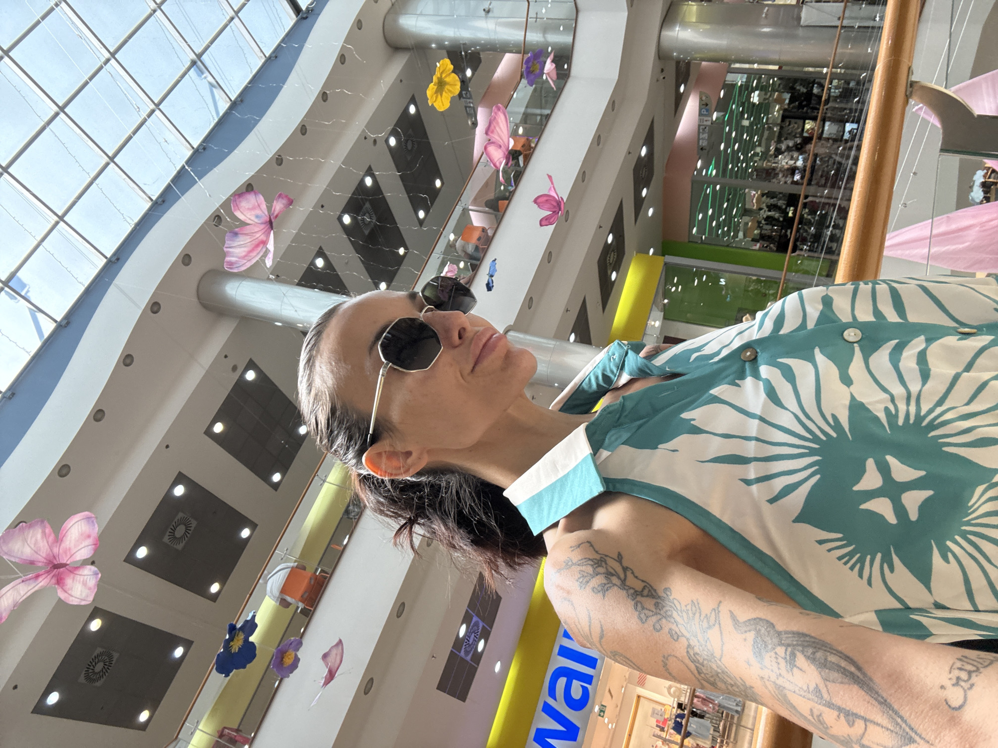 The image shows a woman taking a selfie in a mall. She is wearing sunglasses and a sleeveless white and teal patterned top. Her left arm, visible in the foreground, is covered in tattoos. The background shows a multi-story mall interior with escalators, shops, and decorative hanging butterflies and flowers. The location is identified as Pleven, Bulgaria.
