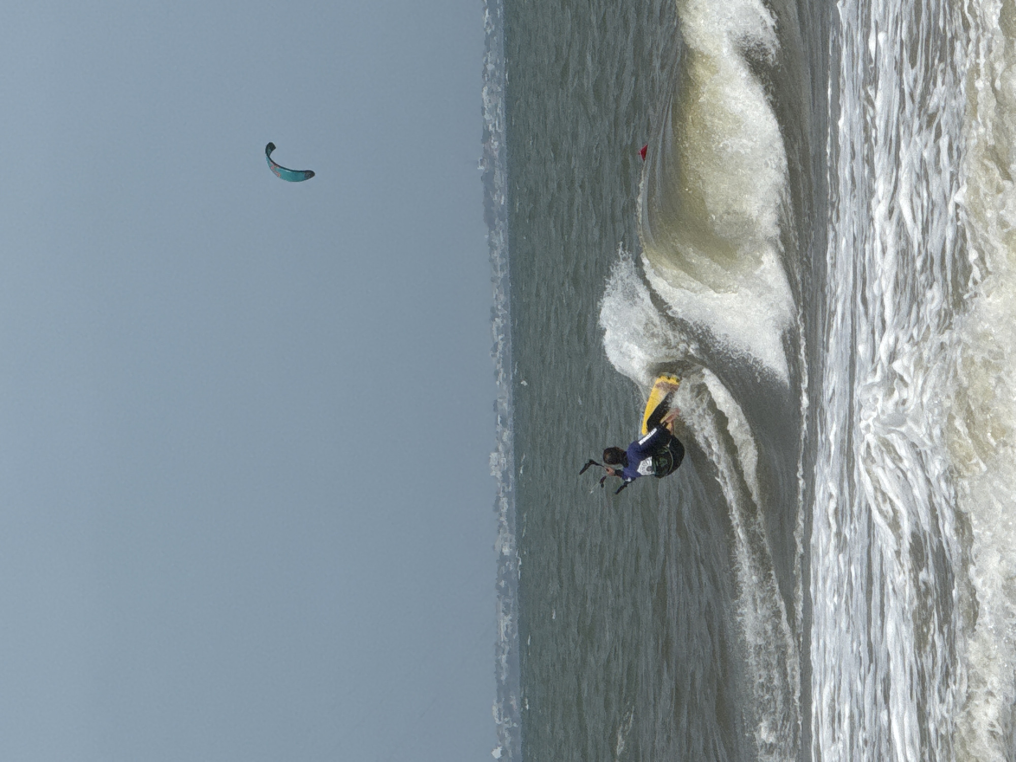 A kitesurfer performs a maneuver on a wave in the ocean, with a distant cityscape visible on the horizon. The kitesurfer, wearing a dark blue suit and a yellow board, is angled towards the viewer and appears to be in mid-air or in the process of turning. The wave is breaking, with white foam and spray around the kitesurfer. In the distance, a hazy line of buildings suggests a city or town. A second, smaller kite is visible in the pale blue sky. The water is choppy and dark gray, and the sky is overcast. This scene could be located along the coast of Phan Thiết, Vietnam, a region known for its beaches and water sports.