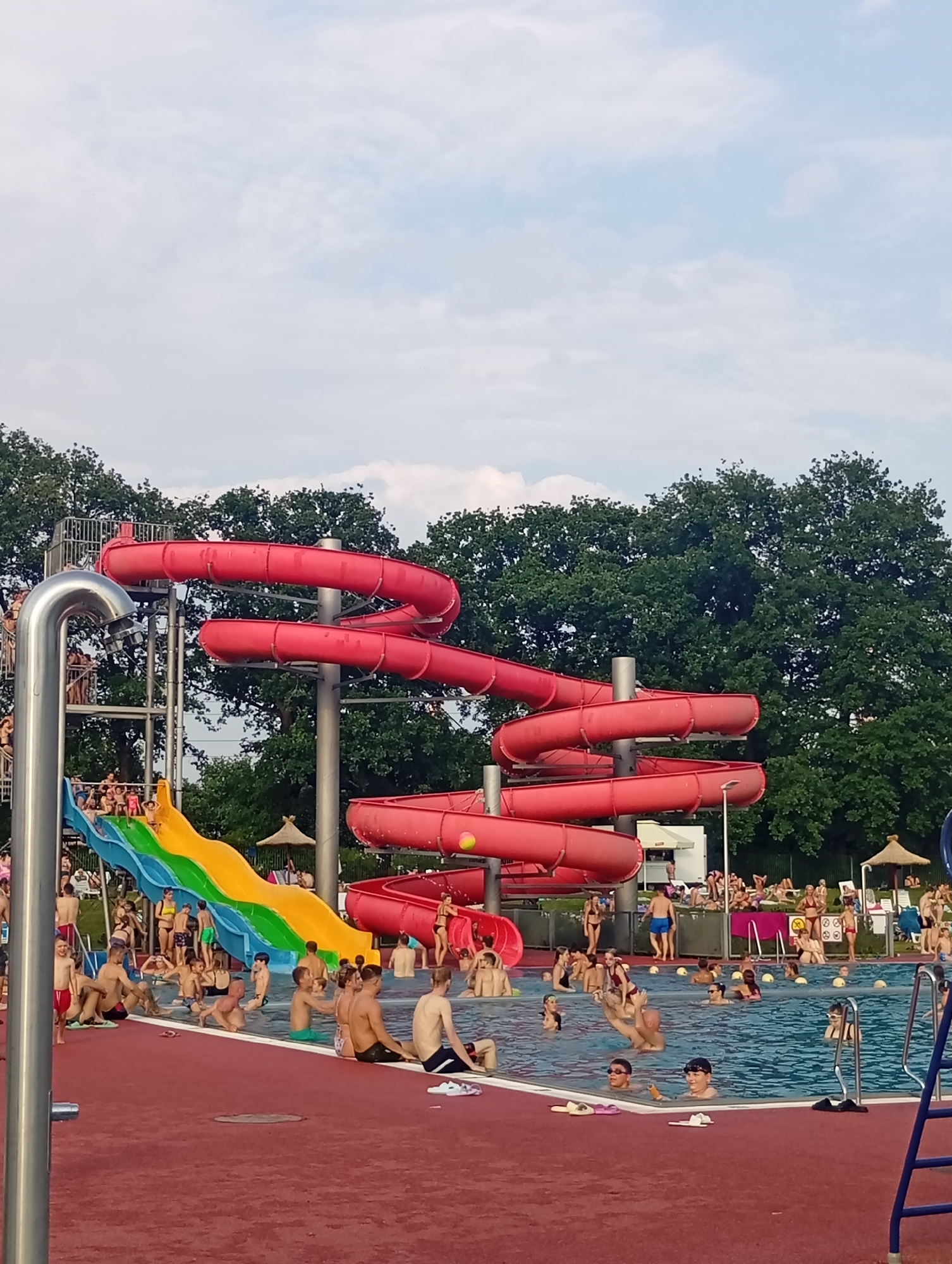 This is a wide-angle, eye-level, outdoor photograph of a crowded public swimming pool area on a cloudy day. The scene is likely in Wrocław, Poland, given the context of a known water park. In the foreground, a red, textured surface, likely a pool deck, surrounds a large blue swimming pool. Numerous people are in the pool, some swimming, others standing or sitting at the edge, and many are partially submerged.
Dominating the background are several large, red spiral water slides. One slide forms a prominent, continuous spiral, while another appears to have a more open, tube-like structure. Adjacent to these slides is a blue and yellow water slide with a green section, also featuring people on it. A metal staircase structure supports the slides.
People of various ages and genders are present, wearing swimwear. Actions include people going down slides, floating in the water, standing in groups, and relaxing. The environment includes lush green trees forming a dense backdrop behind the water park structures. Some lounge chairs and umbrellas are visible further in the background, suggesting areas for sunbathing. The sky is overcast with visible white clouds.