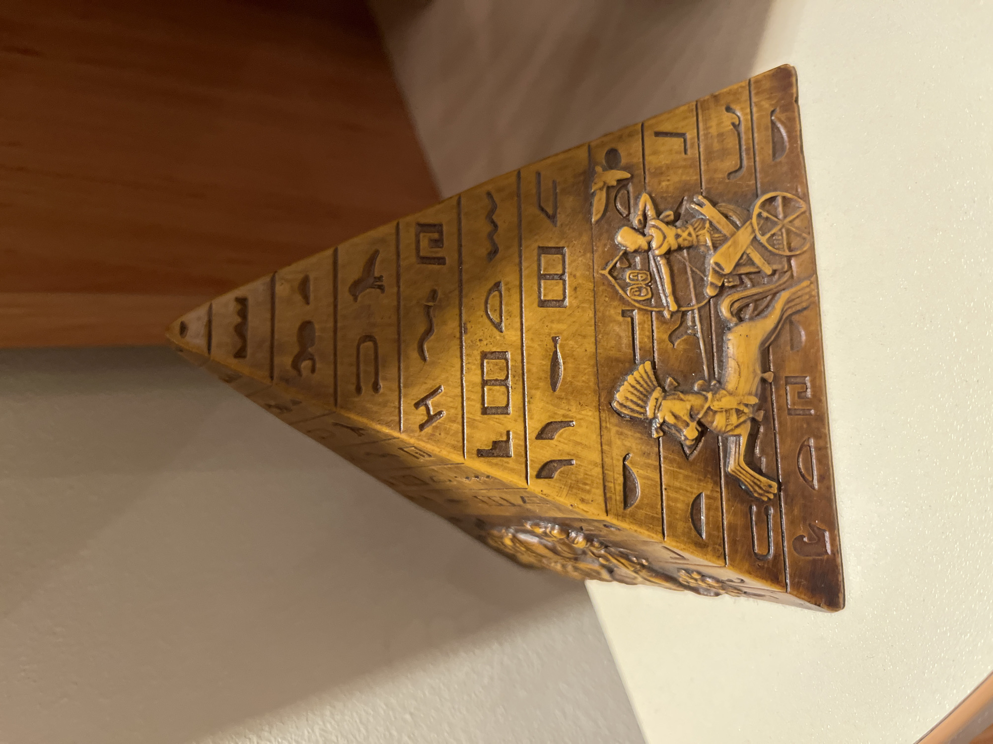 A close-up, eye-level shot shows a golden pyramid-shaped artifact adorned with intricate carvings of hieroglyphs and Egyptian figures, positioned on a flat, pale surface. The background features a light-colored wall with a shadow cast from the left, and a warm, brown, textured surface to the right. One side of the pyramid prominently displays a detailed carving of a chariot pulled by a horse, with an archer aiming a bow and arrow from the chariot. The other visible sides are decorated with rows of carved hieroglyphic symbols and other figures. The lighting appears to be coming from the right, casting subtle shadows on the artifact and the wall.