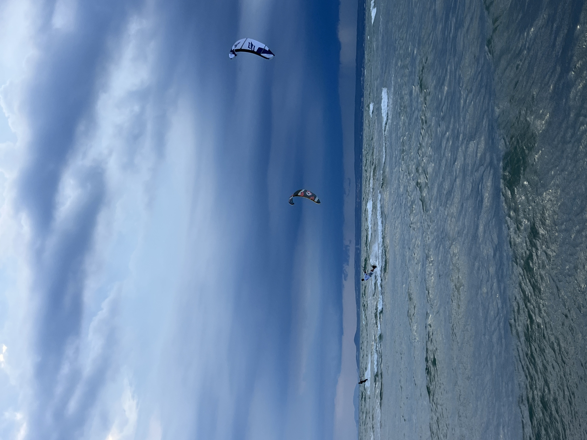 The image captures a dramatic scene of kitesurfing in the waters off Mũi Né, Vietnam, under a turbulent, storm-laden sky. Dark, heavy clouds dominate the upper portion of the frame, with lighter patches suggesting breaks in the storm in the distance. The sea is choppy, with whitecaps indicating strong winds.
Several kitesurfers are visible, scattered across the water. In the middle ground, two kitesurfers are engaged in the activity, their kites forming colorful shapes against the dark sky. To the far left, another kitesurfer is a silhouetted figure against the waves. On the right side of the frame, a larger kitesurfer with a distinctive black and white kite is prominently displayed, appearing to be in mid-air or executing a jump. A second, smaller kitesurfer with a patterned kite is positioned to the left of the larger one.
The horizon line is low, with a faint silhouette of distant land and hills visible. The overall impression is one of powerful natural forces at play, with the kitesurfers skillfully navigating the challenging conditions. The lighting suggests it is either early morning or late afternoon, with the storm clouds casting a dramatic, moody atmosphere over the scene.