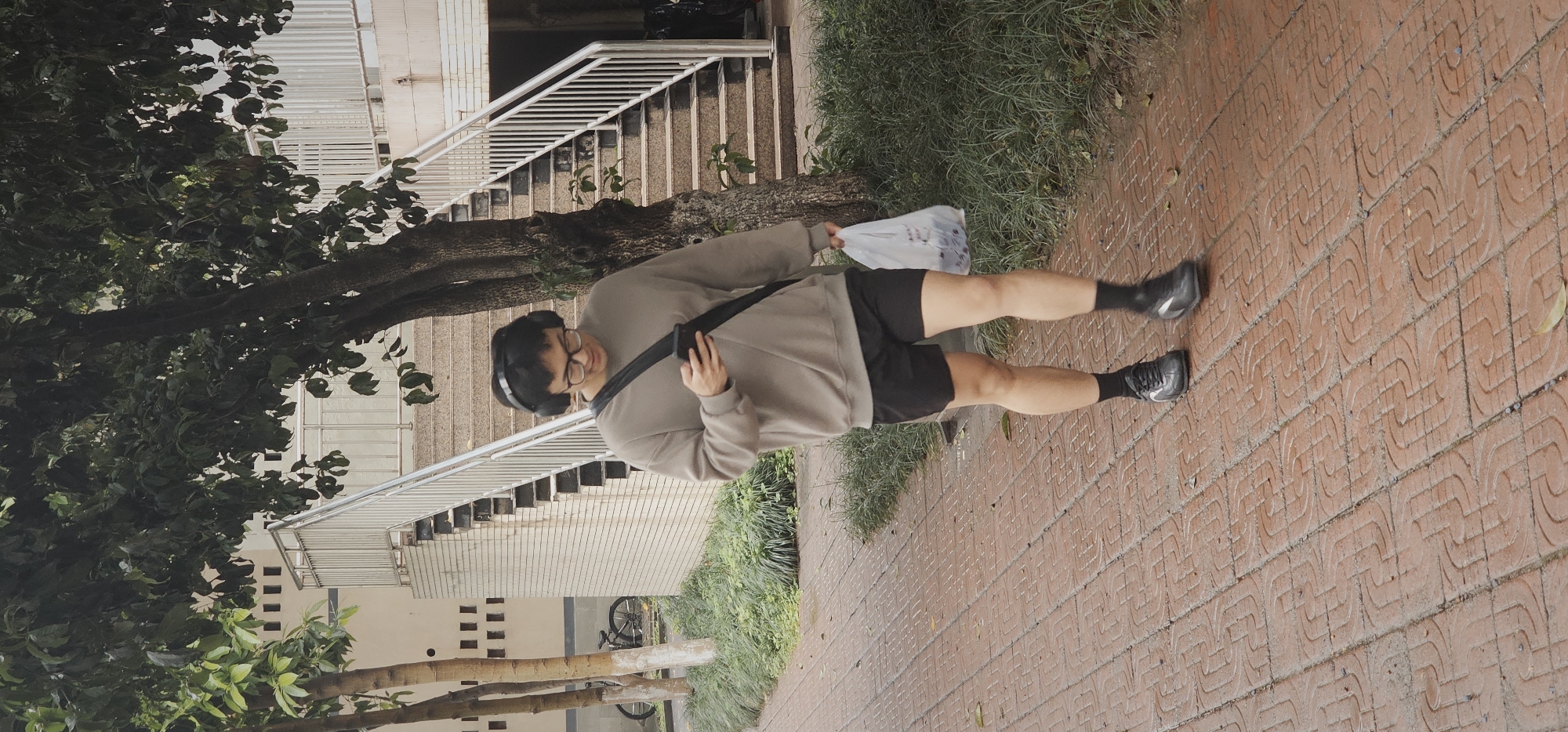 The image shows a man standing on a brick sidewalk next to a patch of green grass. He's wearing a grey sweatshirt, black shorts, black socks, and black shoes. A black strap goes across his body, and he has headphones on his head. He holds a plastic bag in one hand. In the background, there are stairs, trees, and a building. The picture appears to be taken in Yuzhong District, China, because the sidewalks are brick.