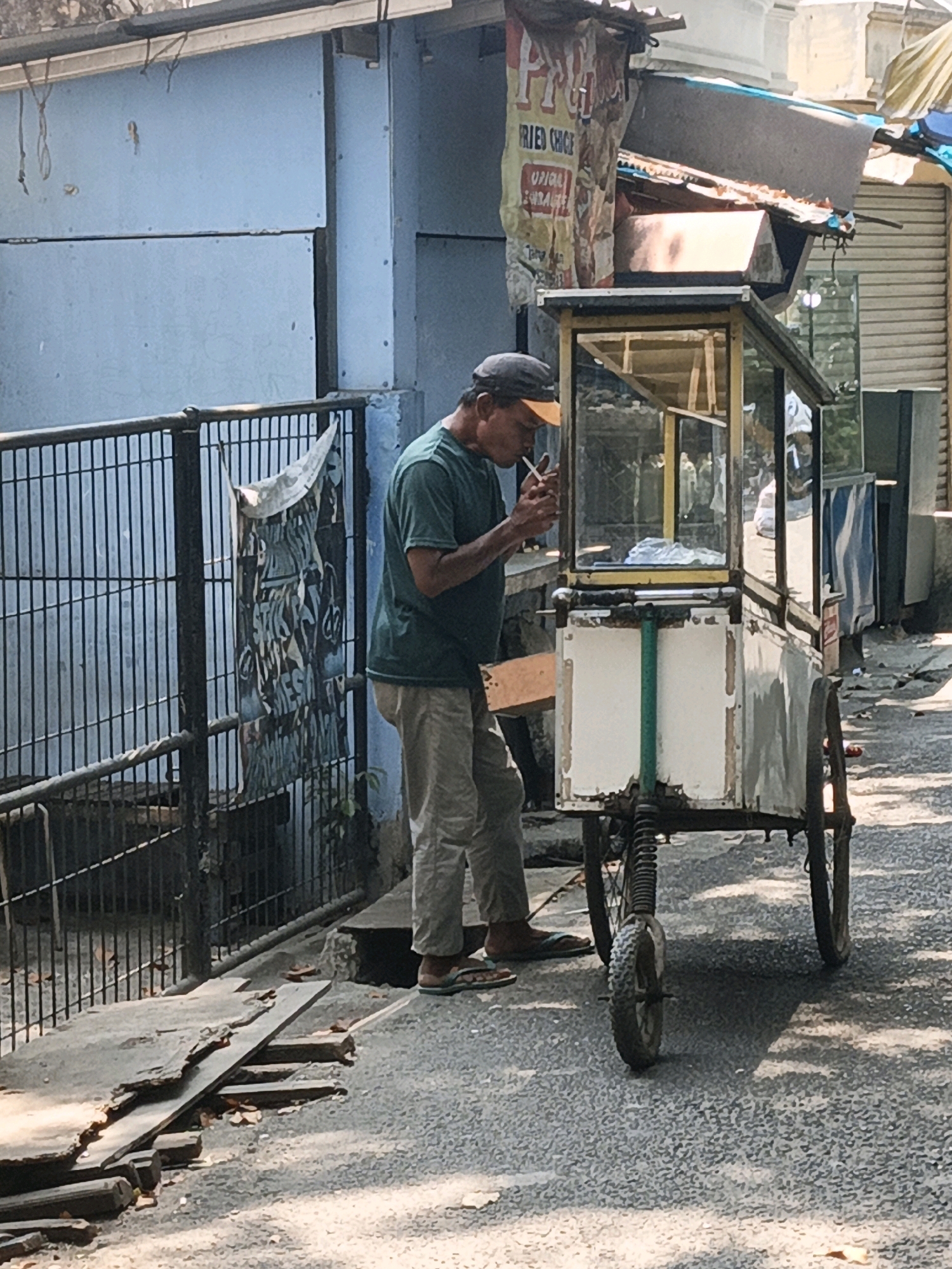 A man stands in front of a food cart on a street in West Jakarta, Indonesia, lighting a cigarette. The man is wearing a green t-shirt, khaki pants, a dark baseball cap with an orange brim, and sandals. He is holding a lit cigarette to his mouth with his right hand, and his left hand is near his face. The food cart is white with a glass display case and a metal frame. It has two wheels on the back and a single wheel in the front. There is a sign on the cart with faded text and images. Behind the man is a blue wall and a black metal fence. The ground is paved with asphalt and there are some fallen leaves and debris. The image is taken in daylight and the sun casts shadows on the ground.