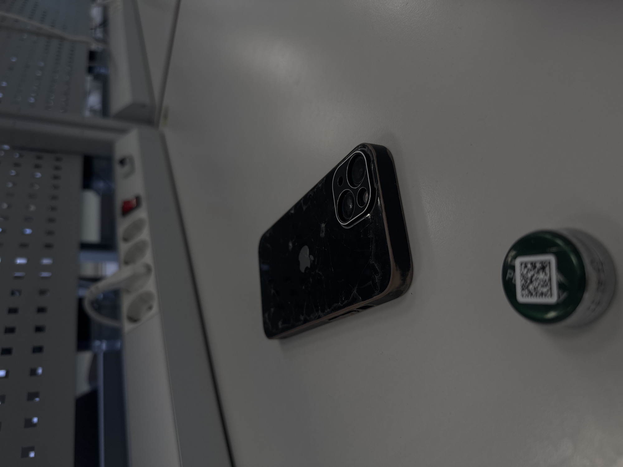 The image shows a black iPhone with a cracked screen attached to a white, seemingly metallic, wall. Beside it, to the right, there is a green cylindrical container with a QR code on top. The phone appears to be attached to the wall in an industrial or commercial setting, possibly a factory, workshop, or server room. There are elements of a rack or panel on the left side, with what looks like power sockets and other electronic components. There is no evidence in the image to indicate that the location is in Perm, Russia. The image contains no people or interactions.
