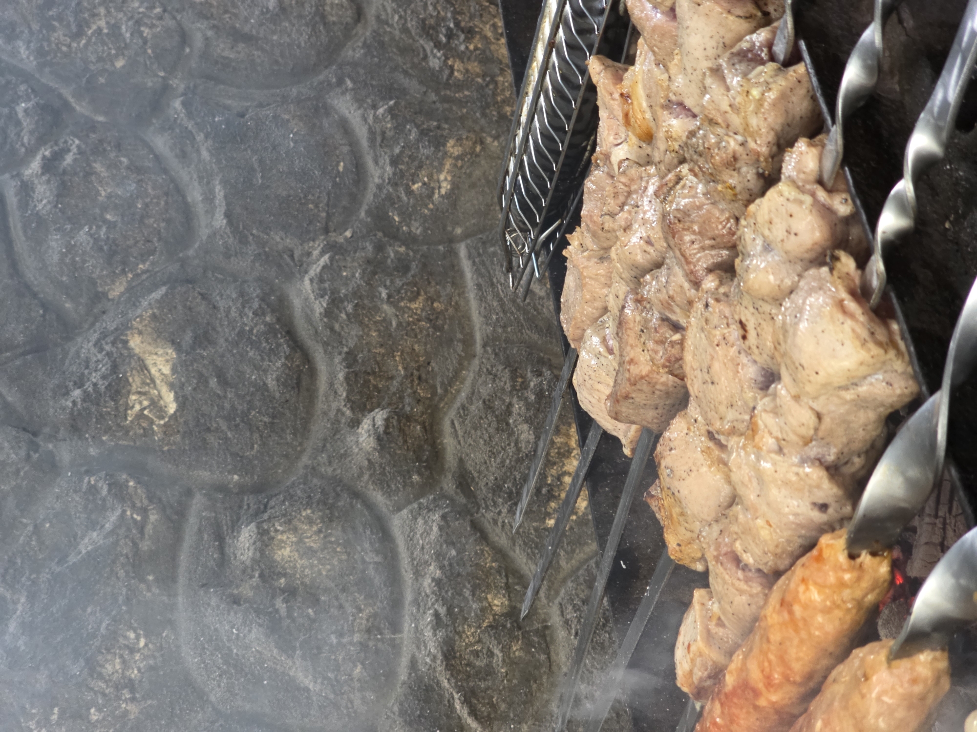 The media file shows a close-up, overhead view of meat being cooked on skewers. The meat is in various stages of cooking, with some pieces appearing more browned and others a lighter pink. The skewers are metal and are arranged vertically, with the meat threaded onto them. The meat is seasoned with black pepper. The skewers are positioned over what appears to be hot coals, as evidenced by the red glow visible beneath the bottom skewer. Wisps of smoke are rising from the meat, indicating that it is actively cooking. The background consists of a rough, textured stone wall. The lighting suggests it is daytime, and there are no indications of weather conditions. The location is identified as Pidhorodne, Ukraine. No people are visible in the frame. The overall scene depicts a barbecue or grilling event, likely outdoors given the stone wall and the cooking method. The focus is on the food and the cooking process.