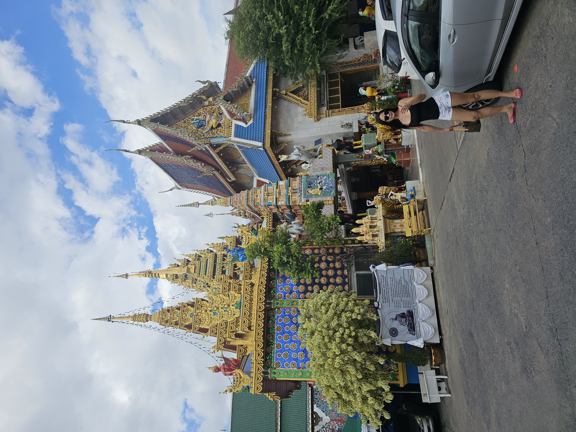 A woman with long dark hair, wearing a black tank top, white shorts with a floral pattern, pink flip-flops, and sunglasses, stands next to a silver car in front of a richly decorated temple in Bangkok, Thailand. She is holding a small brown handbag. The temple features intricate gold ornamentation, colorful statues, and tiered roofs. Behind the woman and to her left, a large banner displays a portrait of a monk and Thai script. Several smaller statues of animals and figures are also visible in front of the temple. The sky is partly cloudy.