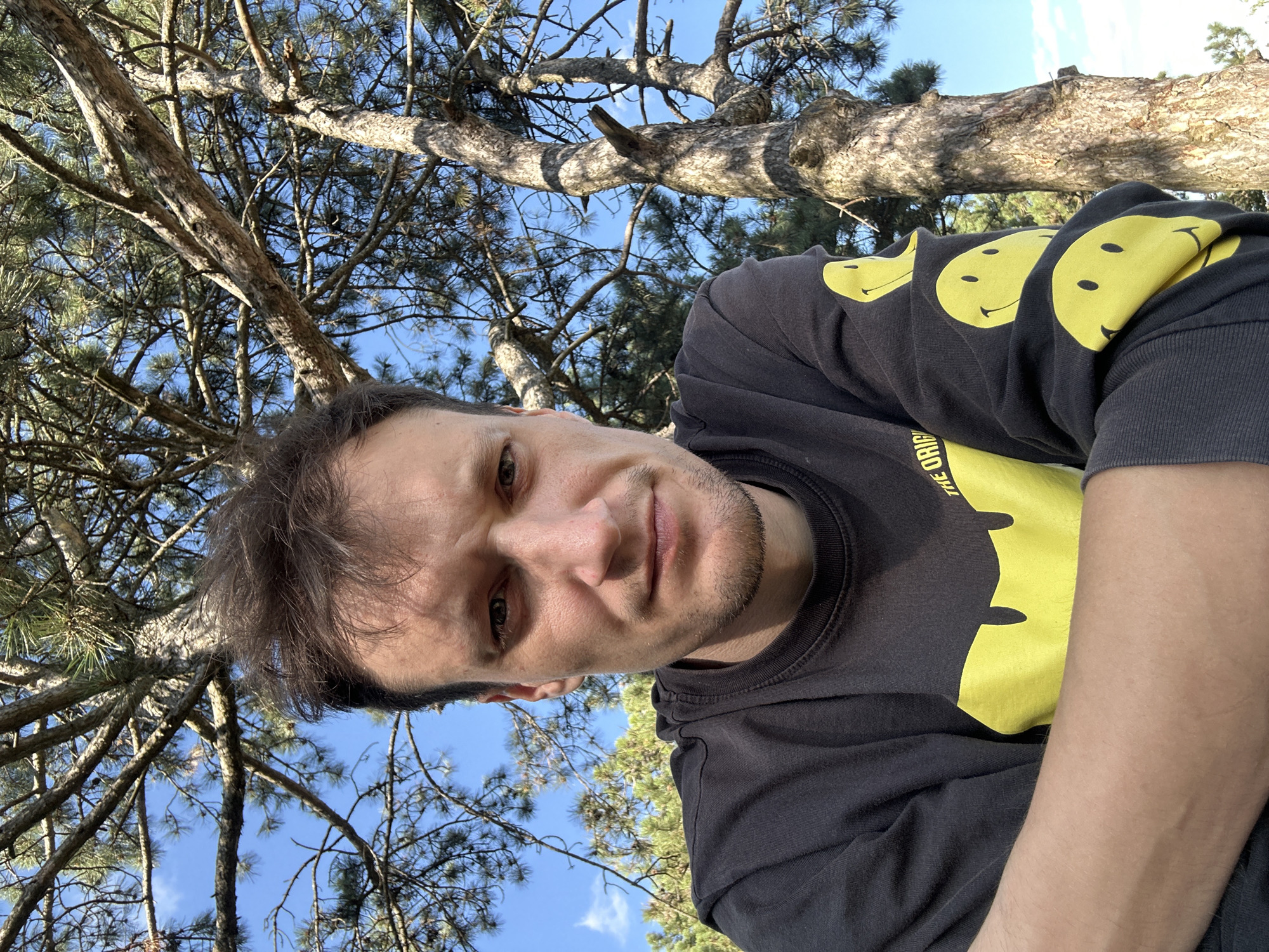 A man is taking a selfie outdoors in front of a tree with a blue sky background. He is wearing a black sweatshirt with yellow smiley faces on the sleeve and a yellow bat symbol on the front. The man has short brown hair and stubble. The tree has rough bark and is partially covered in pine needles. The lighting suggests it is daytime. The location is Makiv, Ukraine.