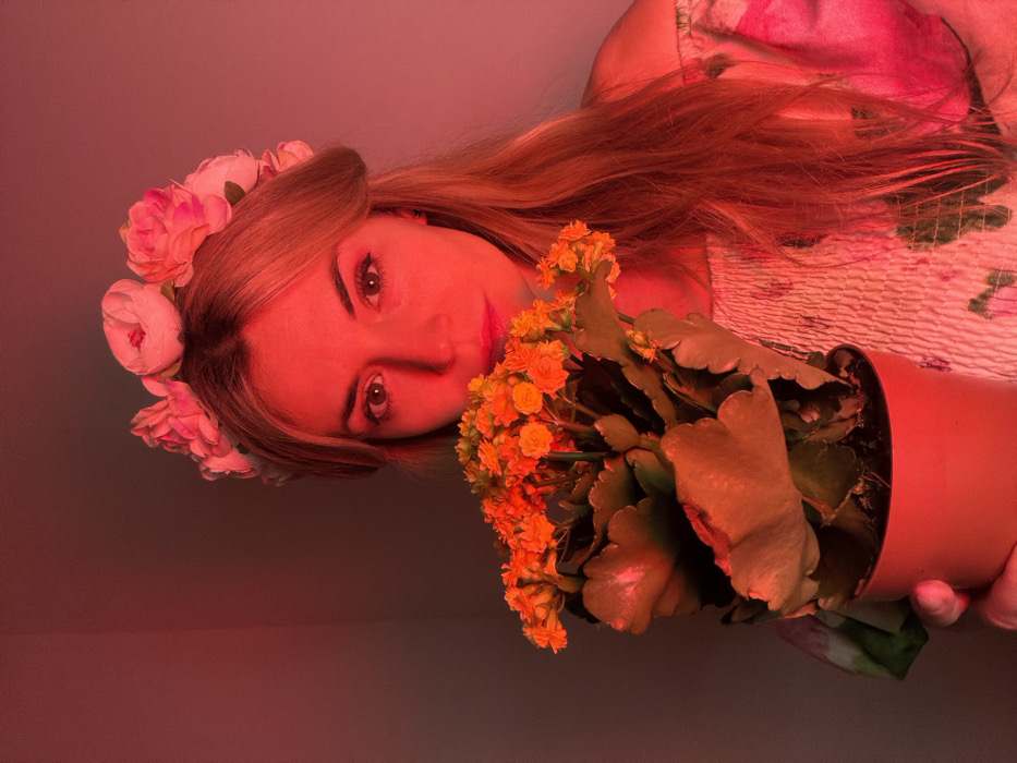 A female subject is depicted from the chest up, centered against a plain, red-lit background. She has fair skin, light brown hair, and maintains direct eye contact with the camera. A floral crown composed of multiple pink and white artificial flowers is worn on her head. Her visible attire consists of an off-the-shoulder, smocked garment with a floral pattern. She holds a potted plant in a terracotta-colored pot with her right hand, visible in the lower right. The plant features numerous small, bright orange flowers and large, dark green, scalloped leaves, positioned to partially obscure the subject's mouth and chin. The entire scene is bathed in a prominent red light, giving the environment and subject a reddish tint. The setting is an interior space, likely a room, located in Budva, Montenegro.