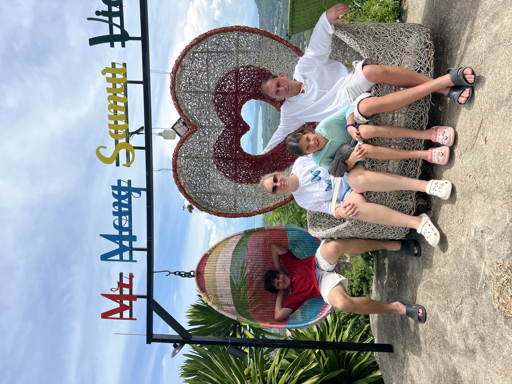 A family of four, two adults and two children, are posing for a photo in front of a large wicker heart sculpture. The family is seated on a wicker sofa. To the left of the sofa, a teenage boy is sitting in a colorful hanging egg chair. Above the family, a sign with colorful letters reads "Mr. Mong Samui Vie." The background shows a lush green landscape with mountains and a body of water, indicative of a scenic viewpoint in Baan Thurian, Thailand. The sky is partly cloudy. The family appears relaxed and happy.
