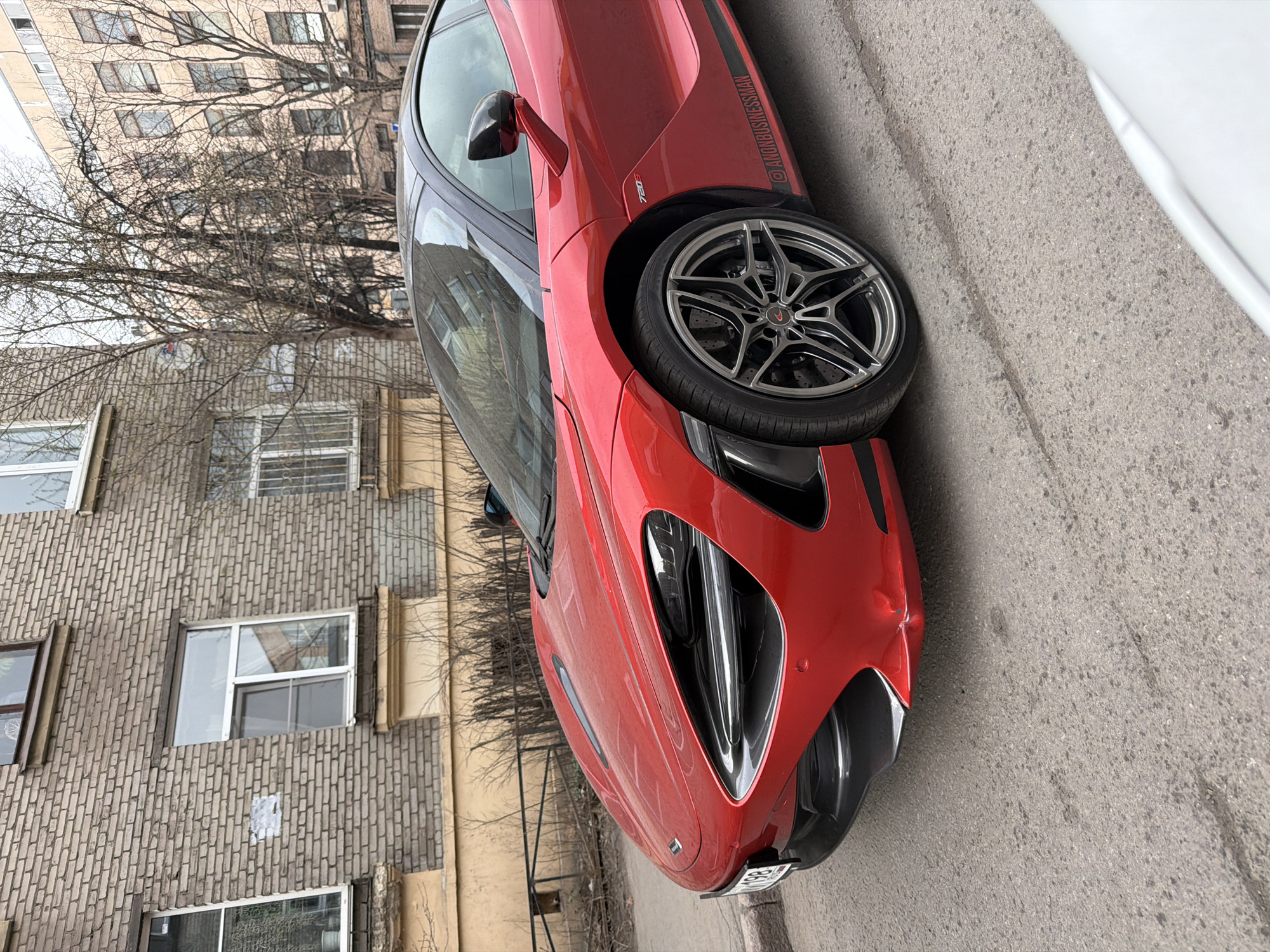 The image shows a red McLaren 720S parked on a street in Saint Petersburg, Russia. The car is positioned next to a building with a textured brick facade and multiple windows. In the background, there is another building and bare trees. The road is paved and shows signs of wear. The car appears to be angled towards the viewer. A portion of a white vehicle is visible in the upper right corner of the image.