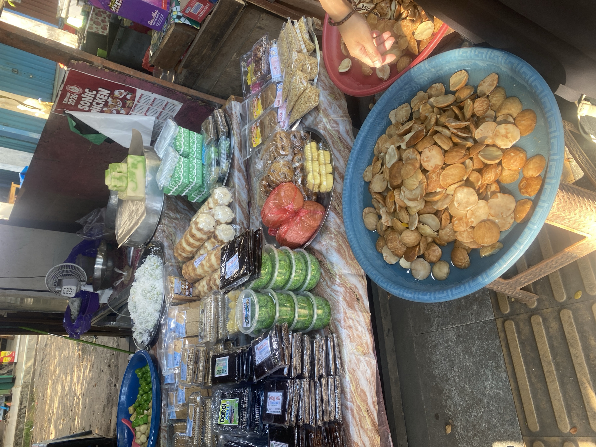 An outdoor market stall in Martapura, Indonesia, displays a variety of food items. The stall features a long table laden with packaged snacks, including what appears to be dodol, cakes, and other sweets. In the foreground, two large blue and red bowls are filled with small, round, light brown nuts or seeds. A person's hand is visible reaching into the red bowl, suggesting a transaction or selection of goods. Behind the table, a fan is positioned near a pile of what looks like shredded coconut. Several colorful plastic containers are stacked, containing green food items. A calendar with the text "Kalender 2026 SONIC CHICKEN" is visible on a reddish-brown vertical surface. The ground is paved with textured tiles. The overall setting suggests a busy street market environment.