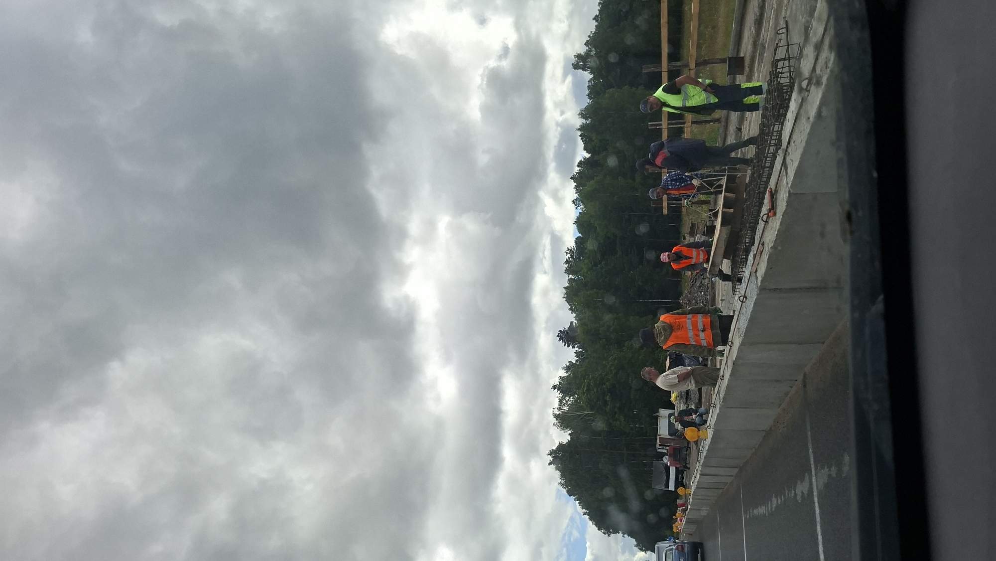 The image shows a construction site on a cloudy day, likely in the Асіпавіцкі сельскі Савет, Belarus, due to the presence of construction workers and a road under development. Several construction workers, some wearing high-visibility vests in orange and green, are engaged in work on what appears to be a road or bridge infrastructure. They are seen interacting with materials, possibly concrete and rebar, suggesting they are in the process of construction. In the background, a dense forest of green trees lines the horizon, with some lighter trucks and vehicles visible near the edge of the construction area. The foreground is dominated by a newly constructed concrete barrier and a paved road. The overall atmosphere is one of ongoing outdoor labor under an overcast sky.