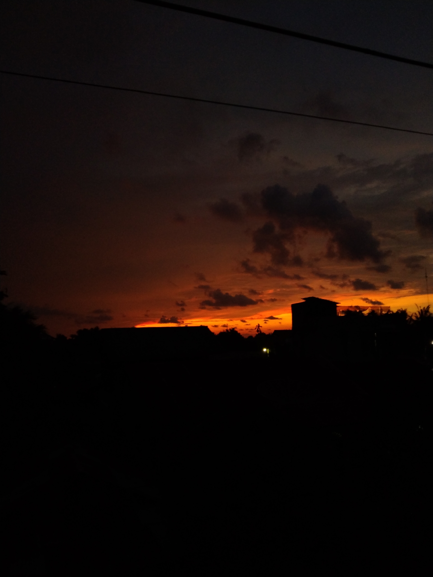 The image depicts a striking sunset or dusk scene, dominated by a vibrant sky and dark silhouettes. The horizon is painted with intense shades of orange and red, indicating the presence of a setting sun. Above this brilliant band, the sky transitions into darker hues of brown and deep gray, with scattered clouds. Some clouds are illuminated by the warm light, appearing orange, while others remain dark against the brighter sky.
In the foreground, the entire landscape is rendered in deep silhouette. The dark outlines of several buildings, including a prominent multi-story structure towards the right, are visible along the horizon line. Dense foliage and trees also form dark masses against the colorful sky. A few small, faint lights can be seen in the distance amidst the dark foreground, possibly indicating artificial lighting from streets or other structures. Two dark, thin lines, likely power lines or utility cables, run horizontally across the upper portion of the frame, passing through the darker part of the sky. No individuals or specific activities are observable. The time of day is clearly sunset or dusk, and the weather appears partially cloudy. There is no visible text in the image. The scene captures the tranquil beauty of twilight over what appears to be a residential or semi-urban area in Bireuenmeunasahdayah, Indonesia.