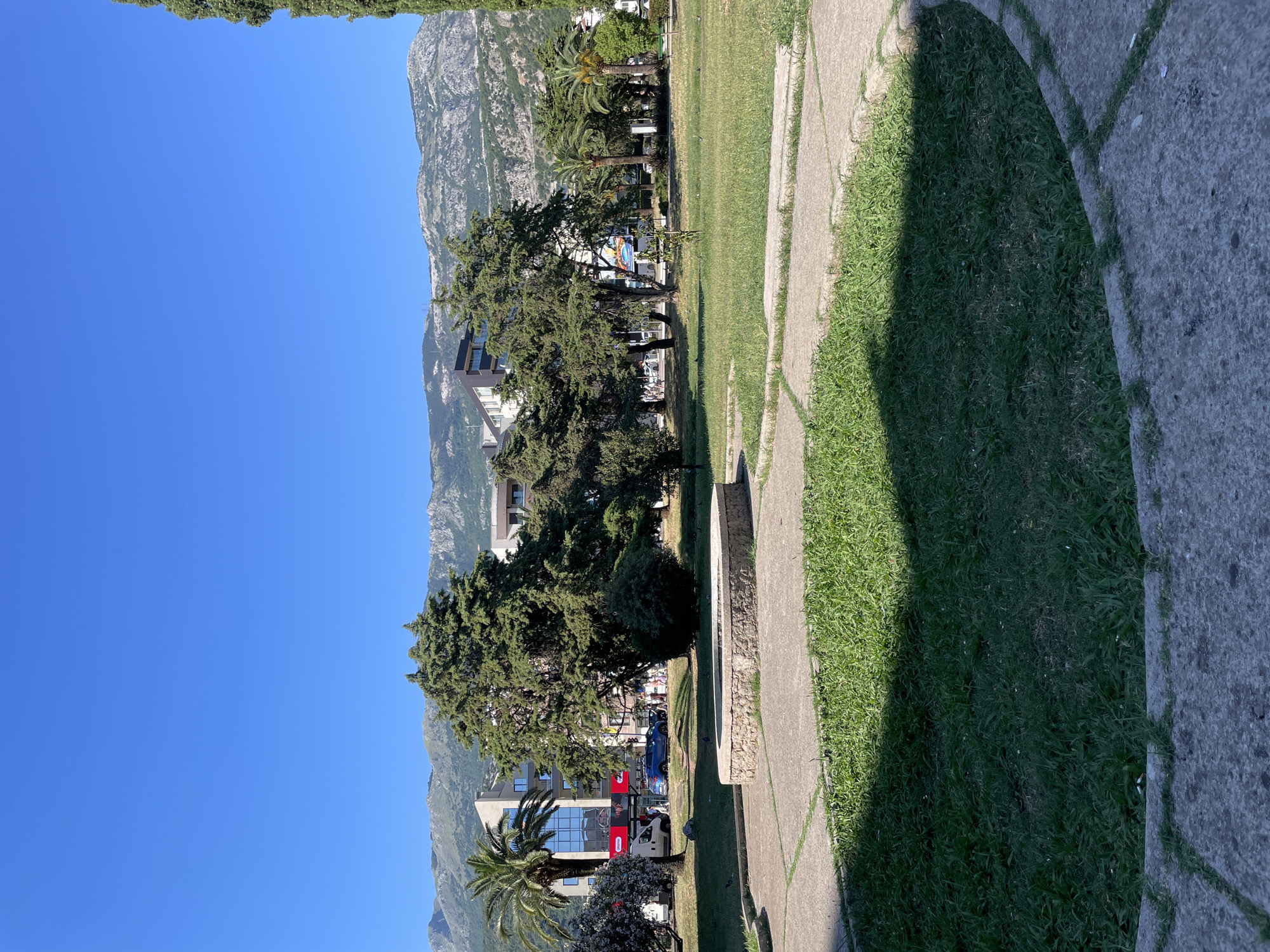 The image captures a view from a park in Budva, Montenegro, on a clear, sunny day. In the foreground, a circular, stone-lined structure, possibly a disused fountain or planter, sits on a paved area surrounded by well-maintained green grass. Paved walkways with a geometric pattern extend across the grass.
In the middle ground, a cluster of mature trees, including palm trees and coniferous trees, provides shade. Behind the trees, modern buildings are visible, some with glass facades, indicating an urban setting. The buildings are situated along a street where a blue car and other vehicles are parked or driving. Several people are walking along the street.
The background is dominated by a range of rugged, gray mountains under a bright blue sky. The overall scene depicts a public space within the city, blending natural elements like trees and grass with urban architecture and a mountainous backdrop.