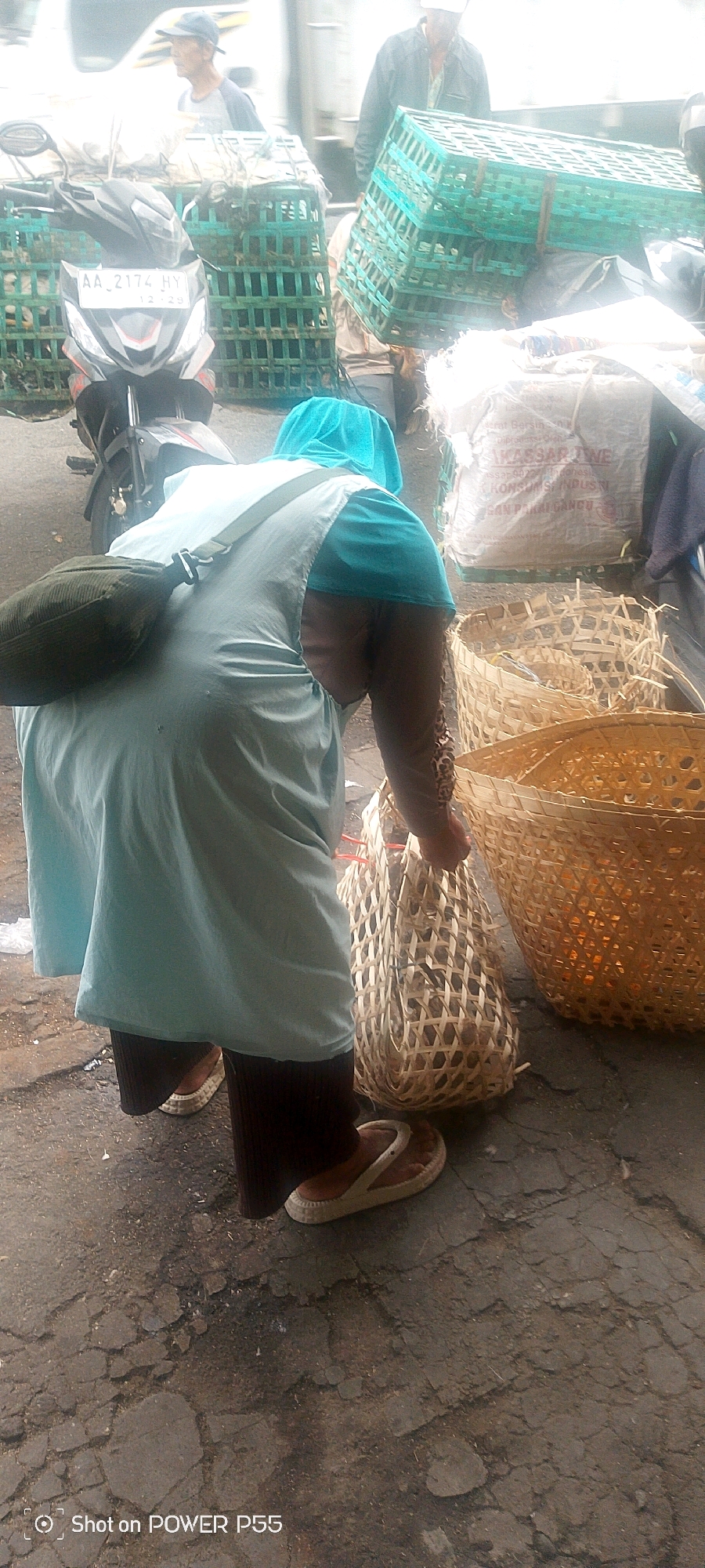 In an outdoor market setting, a person wearing a light blue headscarf and a light blue tunic over dark pants is bent over, holding a woven basket filled with goods. A dark green bag is slung over their left shoulder. Behind them, a motorcycle with a license plate that reads "AA 2174 HY" is parked. In the background, several large green plastic crates and large woven baskets are visible. The ground is paved with cracked asphalt. The scene appears to be in a bustling, possibly agricultural market.