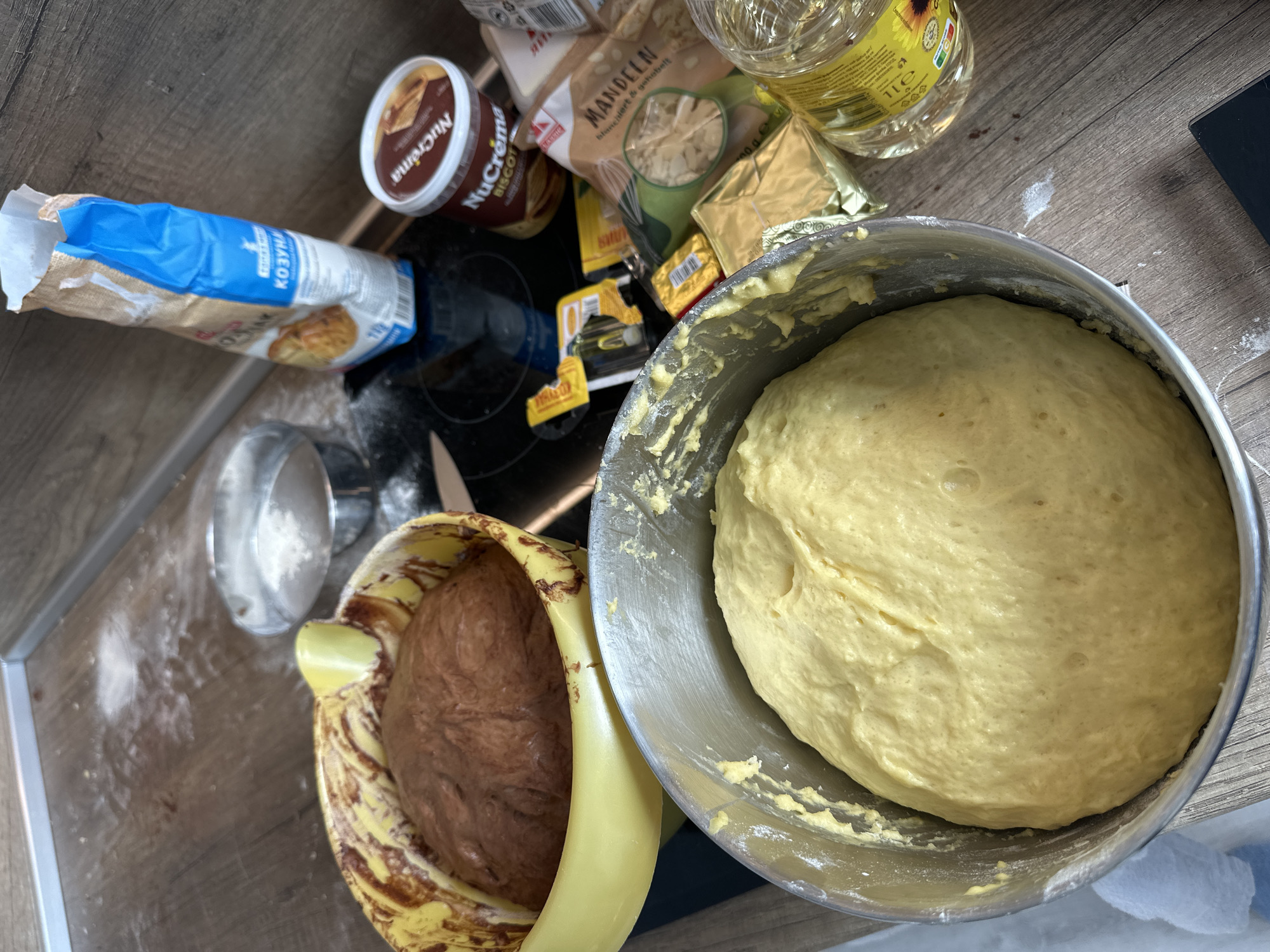 On a wooden countertop, there are two bowls of dough. One bowl, made of metal, contains yellow dough. The other bowl, made of yellow plastic with brown speckles, contains brown dough. Around the bowls, there are various baking ingredients including a bag of flour with the word "КОЗУНАК" on it, a jar of Nucrema Bisco, a bag of sliced almonds, butter, and a bottle of sunflower oil. The scene appears to be indoors, likely in a kitchen. The location is Stara Zagora, Bulgaria.