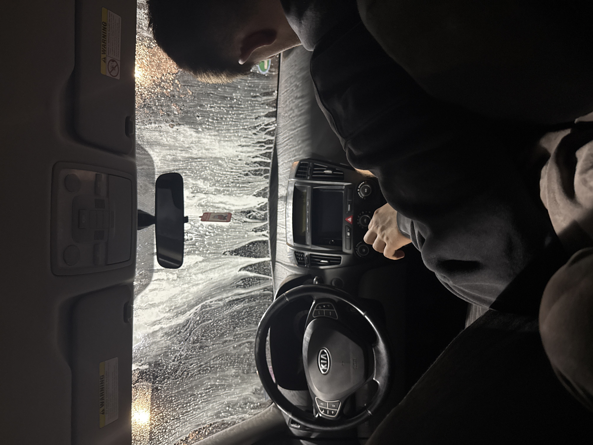 The image depicts the interior of a Kia vehicle undergoing a car wash, as seen from the driver's perspective. Dense foam covers the windshield. A person is partially visible, leaning towards the center console. The steering wheel bears the Kia logo. Other interior elements visible include the rearview mirror, dashboard components, and the car's ceiling. The location within Blagoevgrad, Bulgaria, cannot be determined from the visual information available.