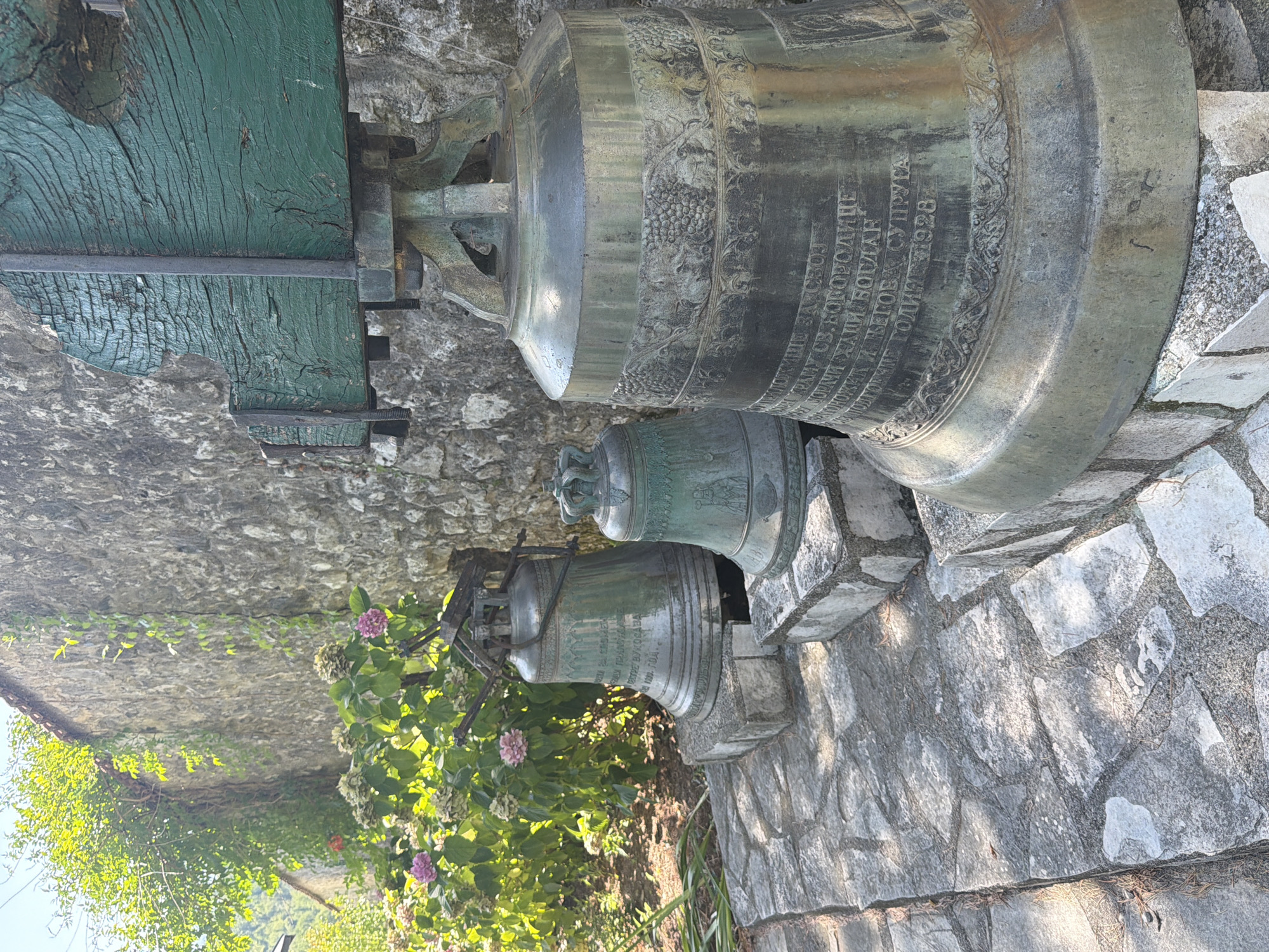 The image depicts three bronze bells arranged in front of a stone wall at Manastir Morača, Montenegro. The bells are of varying sizes, with the largest in the foreground. The largest bell is ornate, featuring inscriptions and decorative bands with grape clusters and foliage. It is supported by a metal structure with a weathered, green-painted wooden beam at the top. The other two bells, smaller and further back, also have inscriptions and decorative elements. They rest on stone bases. Behind the bells, a stone wall is visible, partially covered with ivy and other greenery. Lush vegetation with pink and white flowers is growing to the left of the bells and wall. The ground is paved with irregular grey stones, suggesting an outdoor setting. The scene is bathed in sunlight, casting shadows.