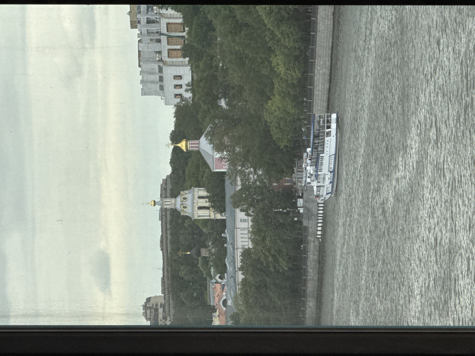 This is a photograph taken through a window overlooking a city scene in Moscow, Russia. In the foreground, a wide body of water, likely a river, occupies the lower half of the image. The water is choppy with visible ripples. A white passenger boat with blue accents is navigating the water, moving from left to right across the frame.
Along the riverbank, there is a stone embankment with a walkway and a line of bollards. Beyond the embankment, a dense line of green trees forms a natural barrier, obscuring the lower parts of the buildings behind them.
Further back, several buildings are visible on a hill. Dominating the skyline are a couple of religious structures with distinctive golden domes. One is a tall, slender bell tower with yellow and white detailing and a golden onion dome. To its left, a smaller dark dome with a cross is visible. To the right, a large, modern-looking white building with many windows and a somewhat unfinished appearance is situated. In the distance, more urban buildings and structures can be seen on the horizon. The sky above is overcast with muted grey and white clouds. The overall lighting suggests a cloudy day.